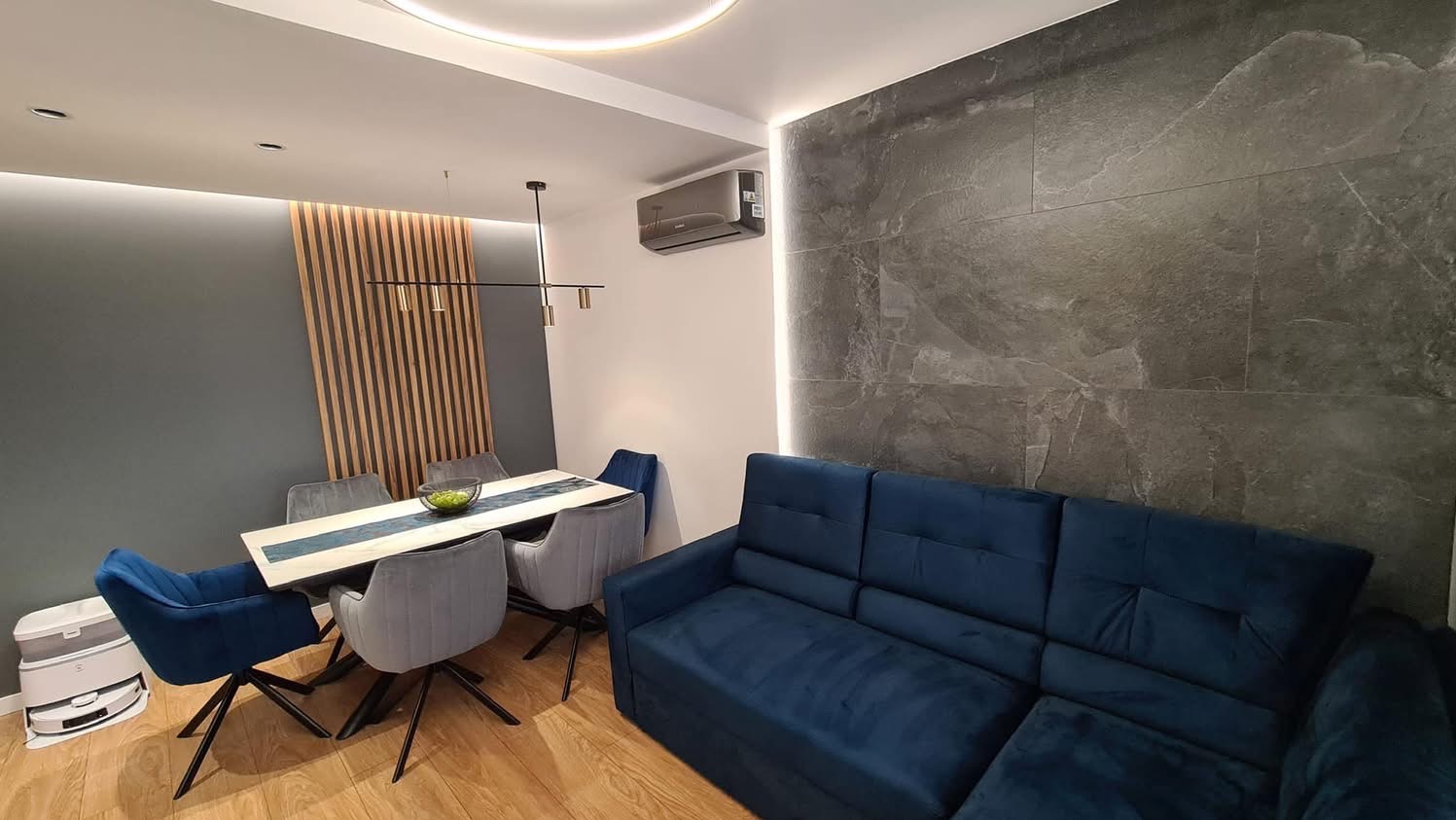 Nowoczesne wnętrze: szara ściana z drewnianymi lamelami, stół z krzesłami, niebieska sofa i ściana z szarego kamienia. Oświetlenie LED podkreśla detale.