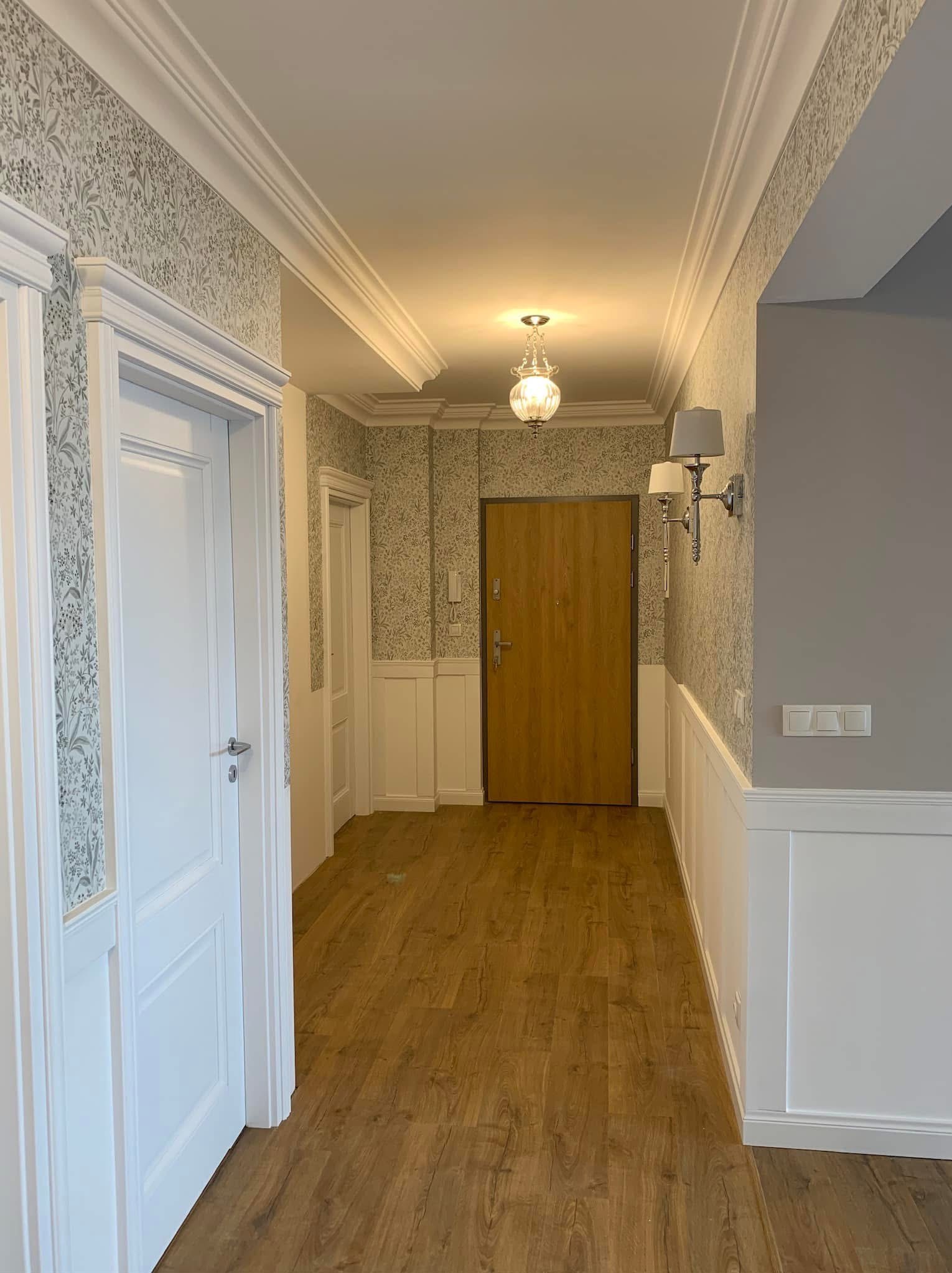 Wykończony korytarz z drewnianą podłogą, tapetą w kwiaty i białymi panelami ściennymi. Widoczne drzwi wejściowe i wewnętrzne, lampy ścienne i sufitowe.