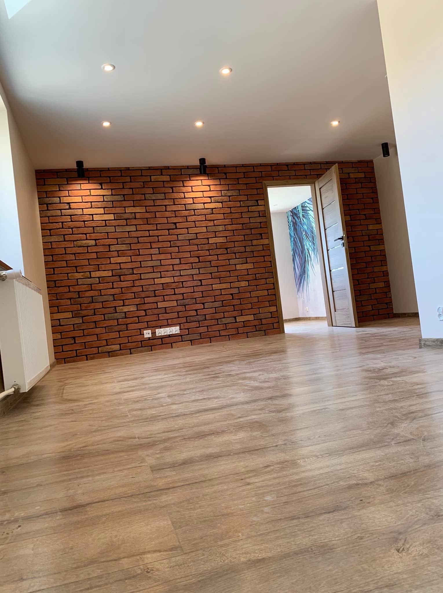 Jasne wnętrze po remoncie: parkiet, ceglana ściana z oświetleniem punktowym, otwarte drzwi z widokiem na tapetę z motywem roślinnym. Nowoczesny design.