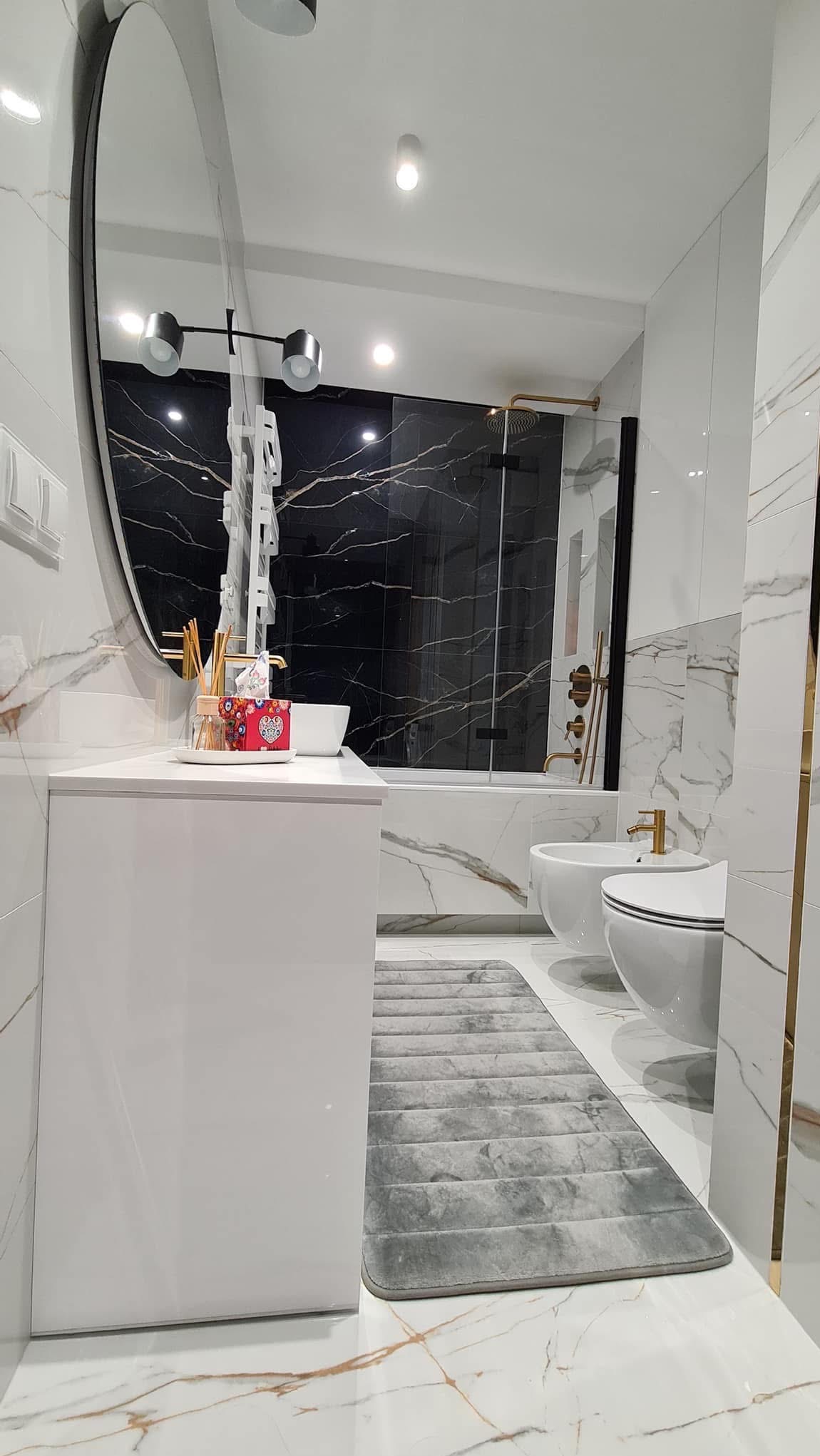 Elegancka łazienka z czarno-białymi marmurowymi płytkami, owalnym lustrem i szarą matą podłogową. Widoczne białe sanitariaty i złote akcenty.