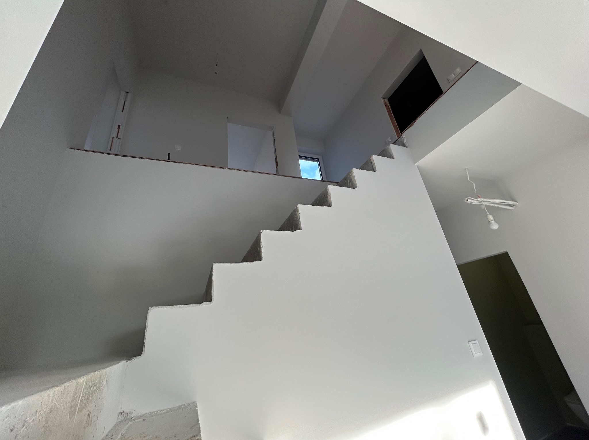Surowe schody betonowe w jasnym wnętrzu, widok z dołu. Świeżo pomalowane ściany, widoczne otwory drzwiowe i okno. Minimalistyczny design w trakcie wykończenia.
