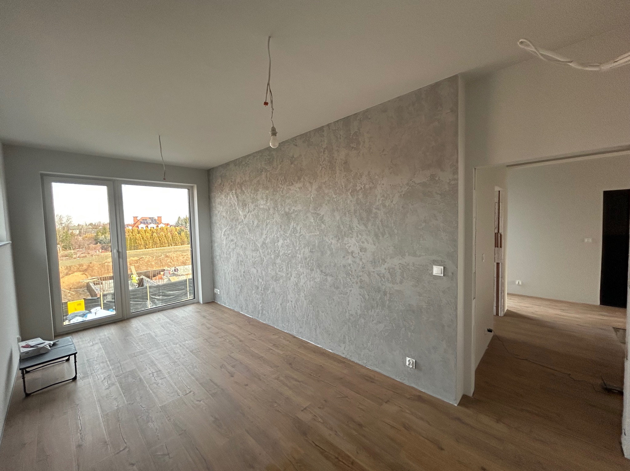 Pokój po wykończeniu: parkiet, ściana z efektem dekoracyjnym, duże okno z widokiem na budowę. W tle widoczne kolejne pomieszczenia.