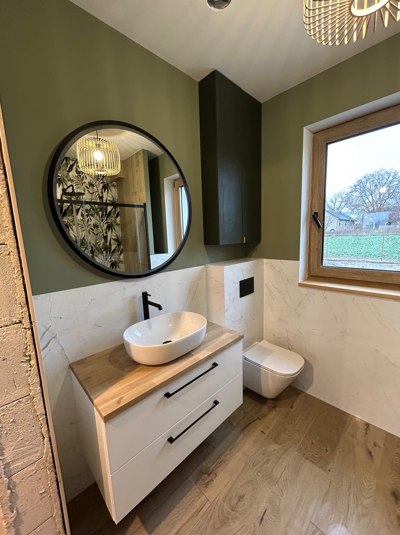 Nowoczesna łazienka z zielonymi ścianami, okrągłym lustrem i drewnianymi akcentami. Umywalka nablatowa na szafce z szufladami, toaleta podwieszana.