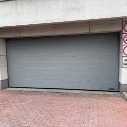 PM Tech Systemy Automatyki Bram Paweł Mirkowicz - Szara segmentowa brama garażowa z poziomymi panelami, zamontowana w nowoczesnym budynku. Widoczne znaki drogowe i sygnalizacja świetlna.