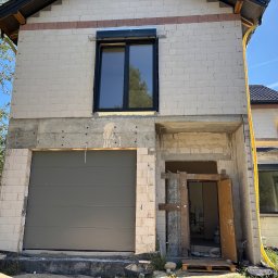 PM Tech Systemy Automatyki Bram Paweł Mirkowicz - Szara brama garażowa segmentowa w surowym stanie budynku z betonu komórkowego. Widoczne okno z roletą zewnętrzną i żółty peszel. Budowa w toku, bez wykończenia.