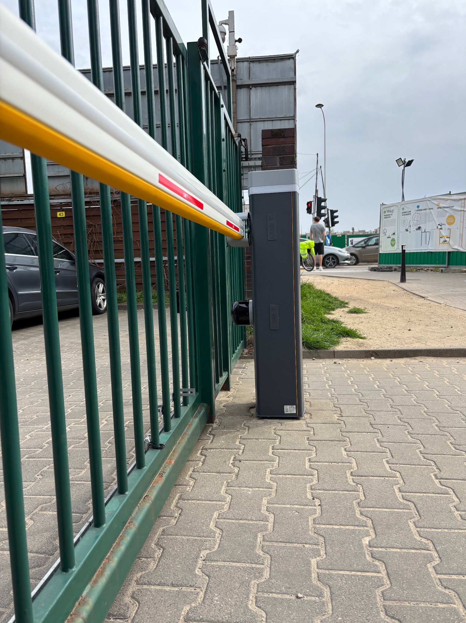 Szlaban automatyczny z zielonymi prętami ogrodzenia, widok z dołu na tle ulicy i przechodnia. Słupek szlabanu szary, z żółto-białym ramieniem. Kostka brukowa na pierwszym planie.