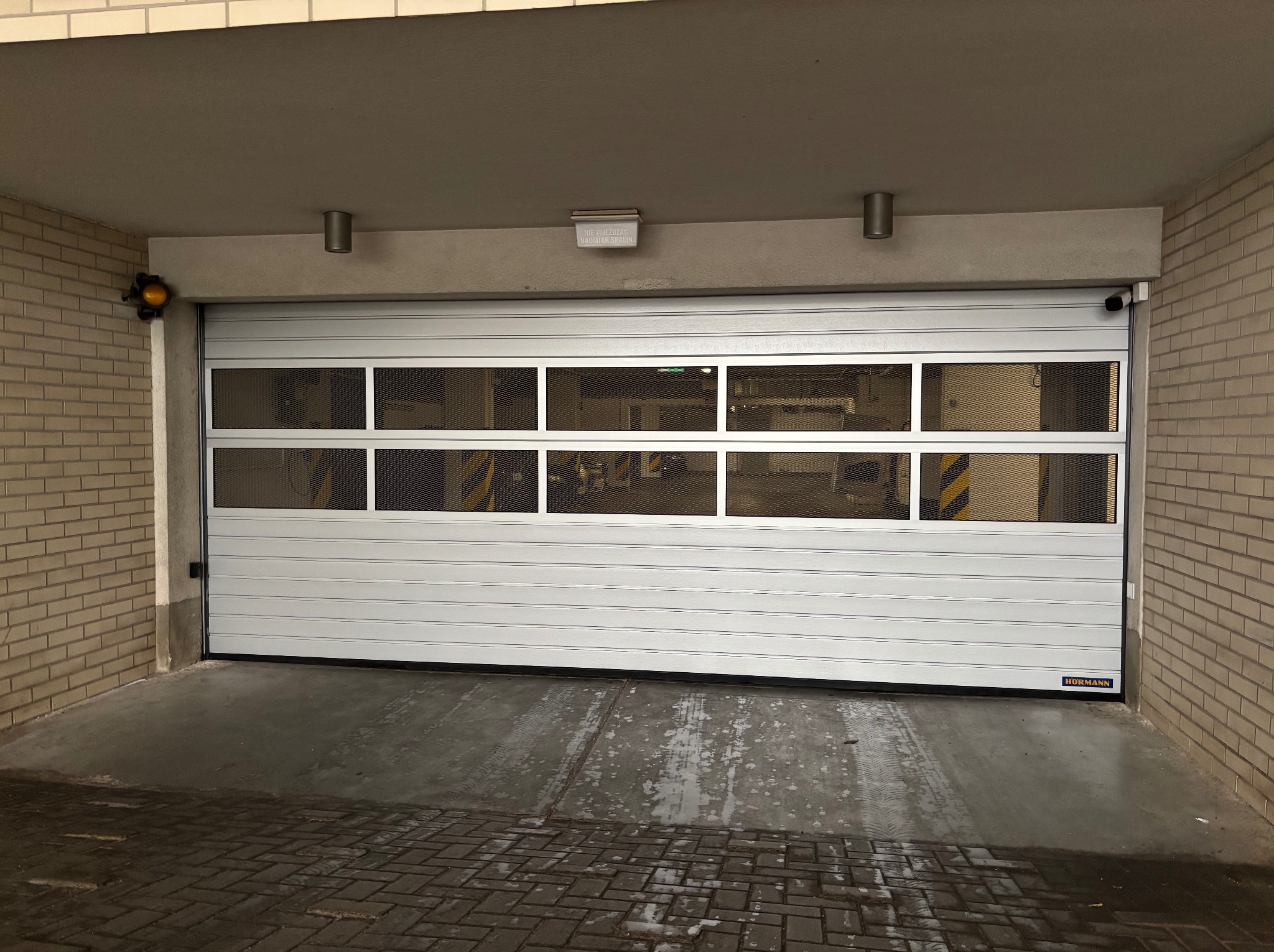 Zamknięta, segmentowa brama garażowa Hormann z poziomymi panelami i przeszkleniami z siatki, zamontowana w garażu z ceglanymi ścianami i betonową posadzką.
