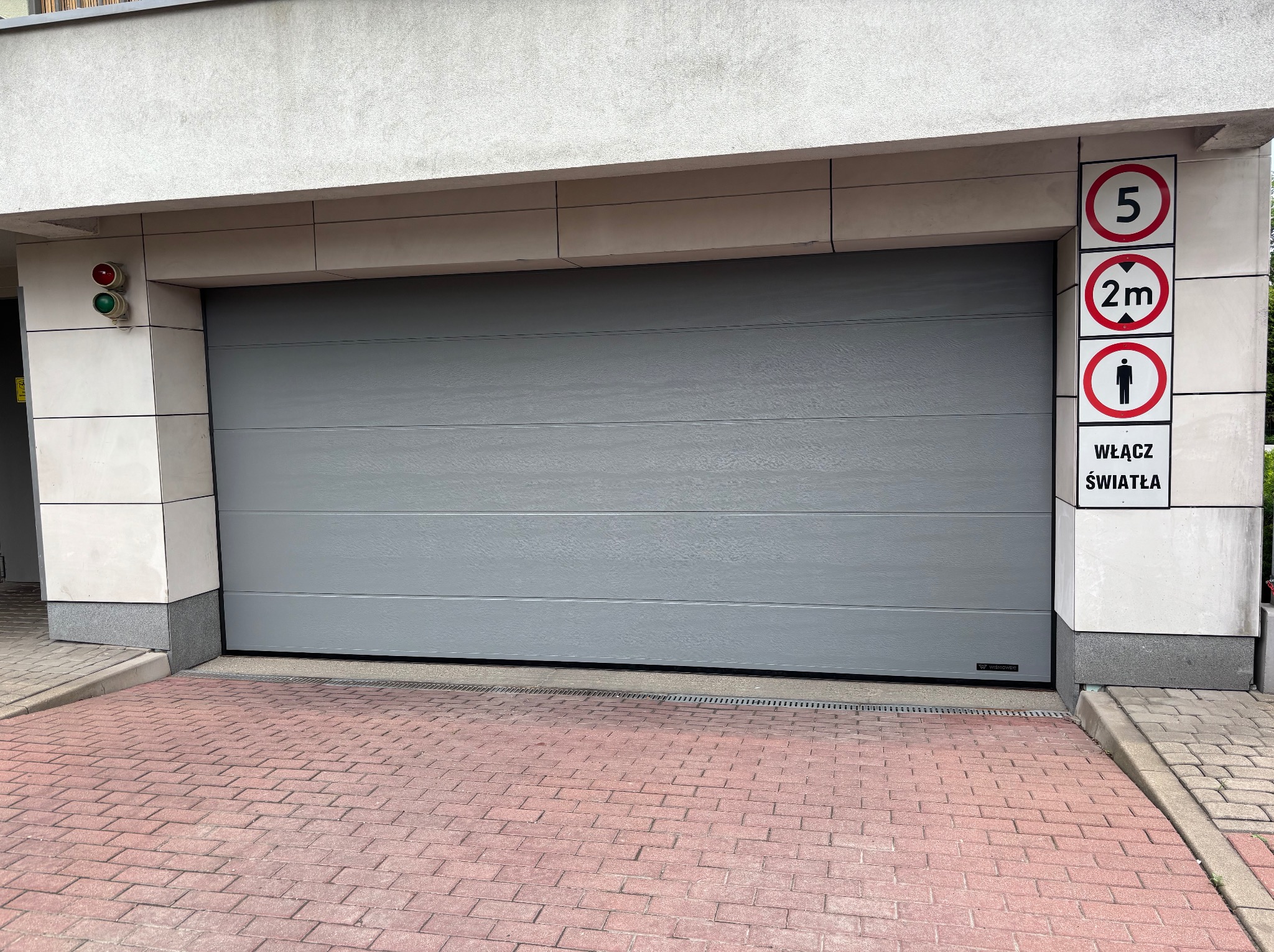 Szara segmentowa brama garażowa z poziomymi panelami, zamontowana w nowoczesnym budynku. Widoczne znaki drogowe i sygnalizacja świetlna.