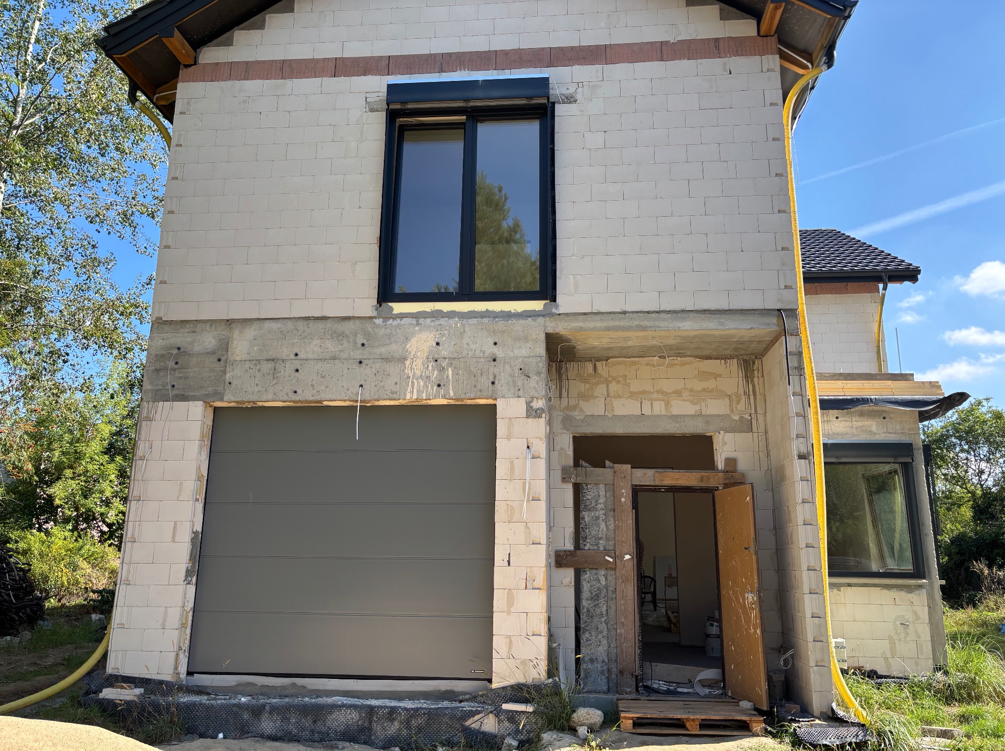 Szara brama garażowa segmentowa w surowym stanie budynku z betonu komórkowego. Widoczne okno z roletą zewnętrzną i żółty peszel. Budowa w toku, bez wykończenia.