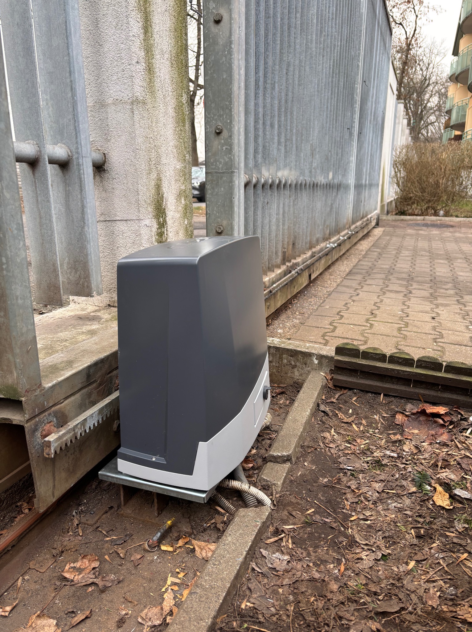 Szary automat do bramy przesuwnej z zębatką, zamontowany w Warszawie. Widoczne przewody i liście na ziemi. Metalowa brama w tle.