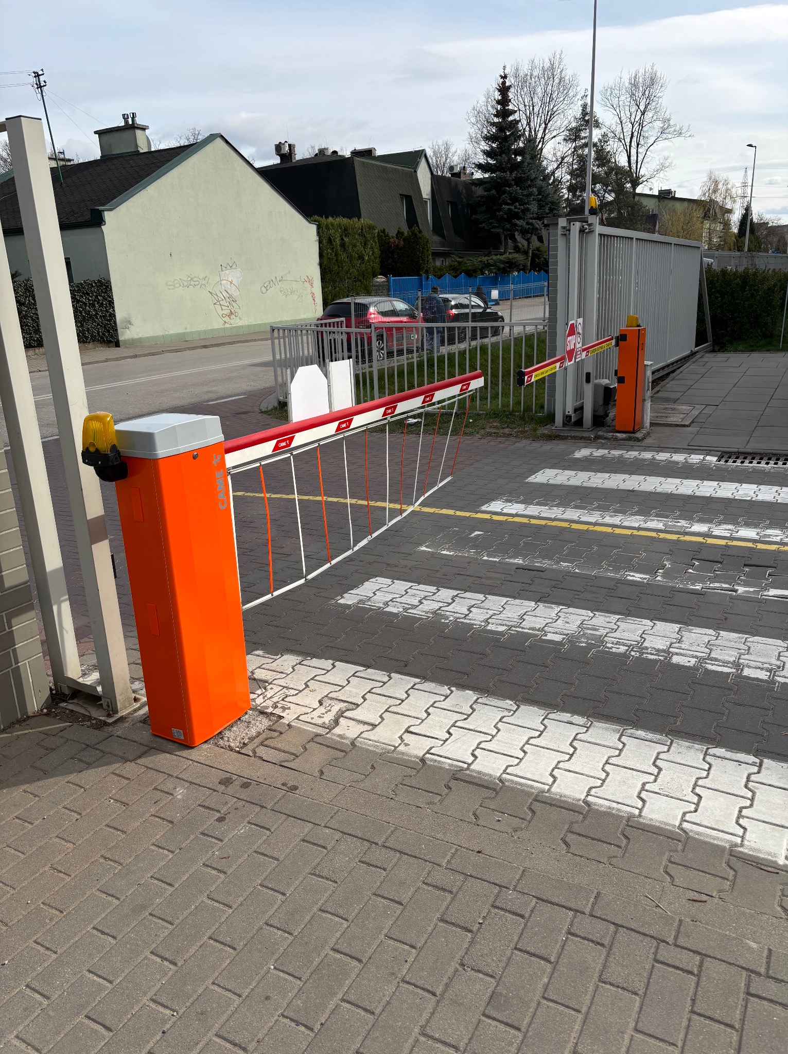 Pomarańczowy szlaban automatyczny CAME z białymi i czerwonymi poprzeczkami, widoczny na tle szarej kostki brukowej i budynków. Oświetlenie dzienne.