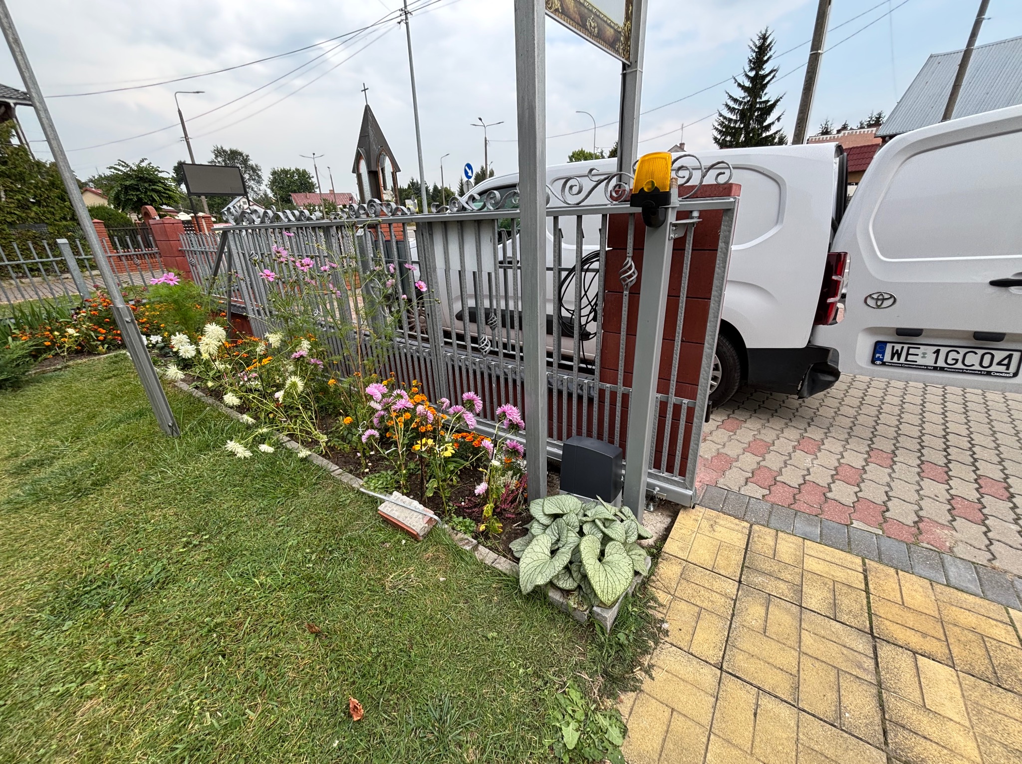 Metalowa brama wjazdowa z automatyką, widoczna lampa ostrzegawcza i sterownik. W tle zaparkowany biały samochód dostawczy i fragment budynku.