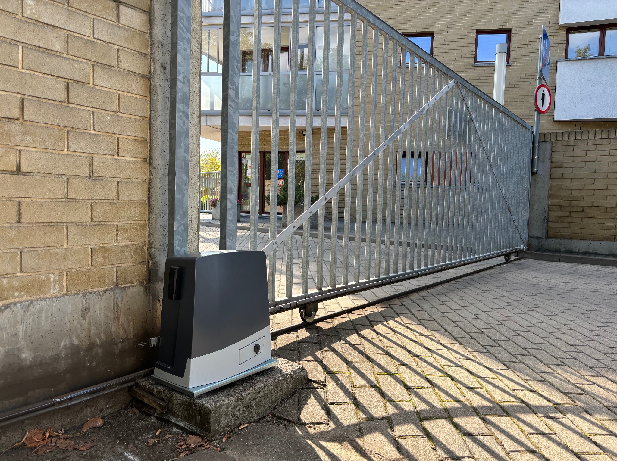 Otwarta, przesuwna brama wjazdowa z szarego metalu, z widocznym napędem automatycznym, zamontowana na betonowym fundamencie. W tle budynek mieszkalny.