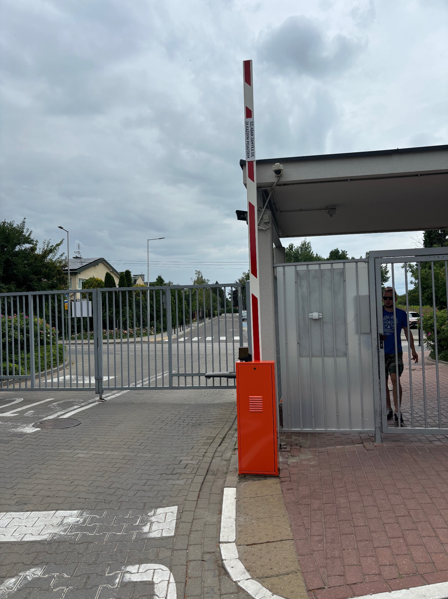Szara brama wjazdowa z automatyczną szlabanem w kolorze pomarańczowym, widok na ulicę i przechodzącego mężczyznę. Słoneczny dzień, lekko pochmurne niebo.