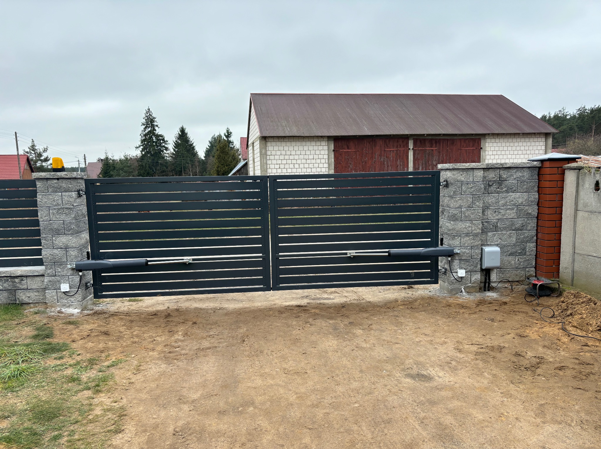 Nowoczesna, pozioma brama wjazdowa z automatyką, zamontowana na murowanych słupkach. Widoczny siłownik i skrzynka sterująca. Tło: budynek gospodarczy i drzewa.