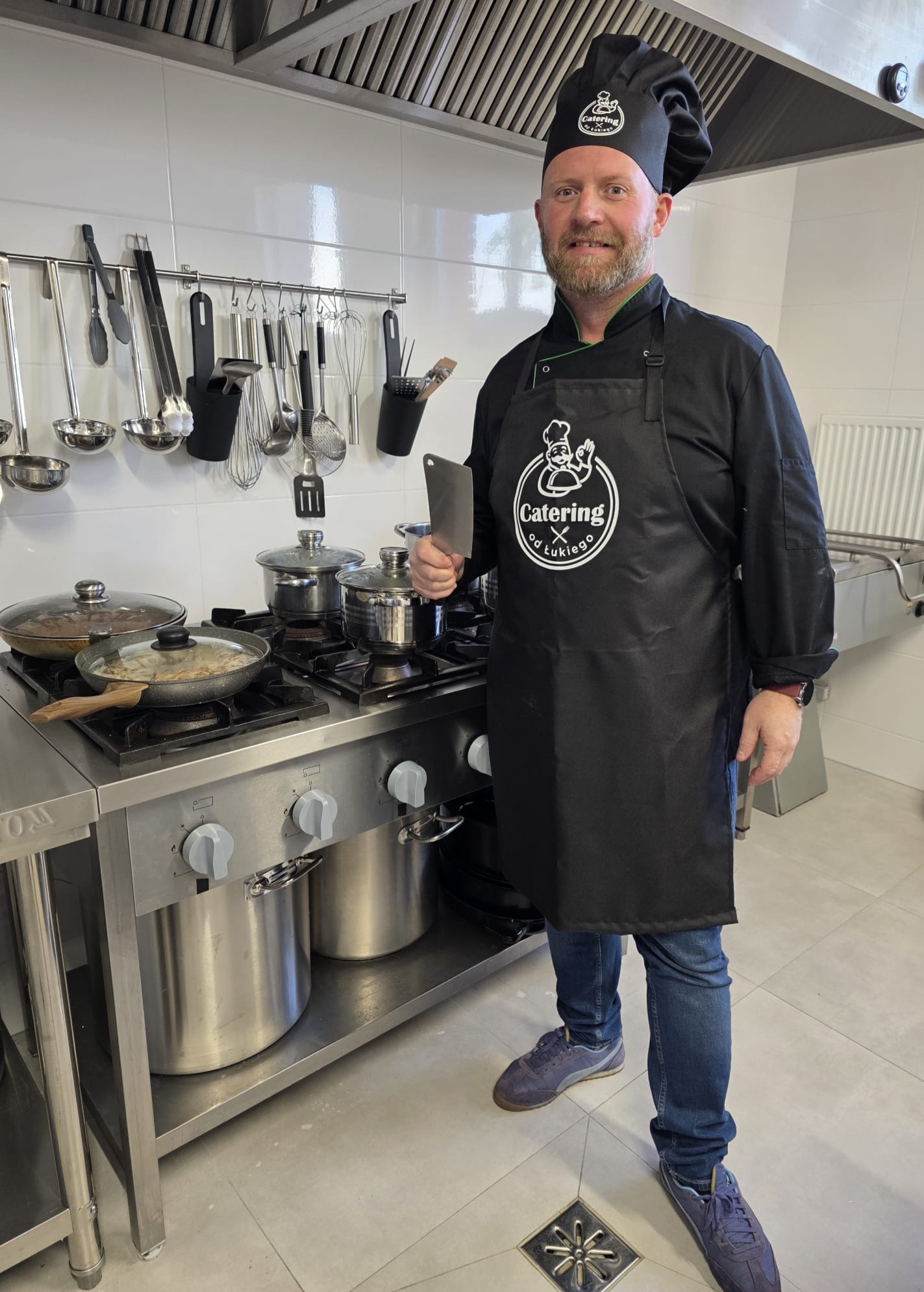 Uśmiechnięty kucharz w czarnym fartuchu z logo 'Catering od Łukiego' trzyma tasak na tle profesjonalnej kuchni z garnkami na kuchence.