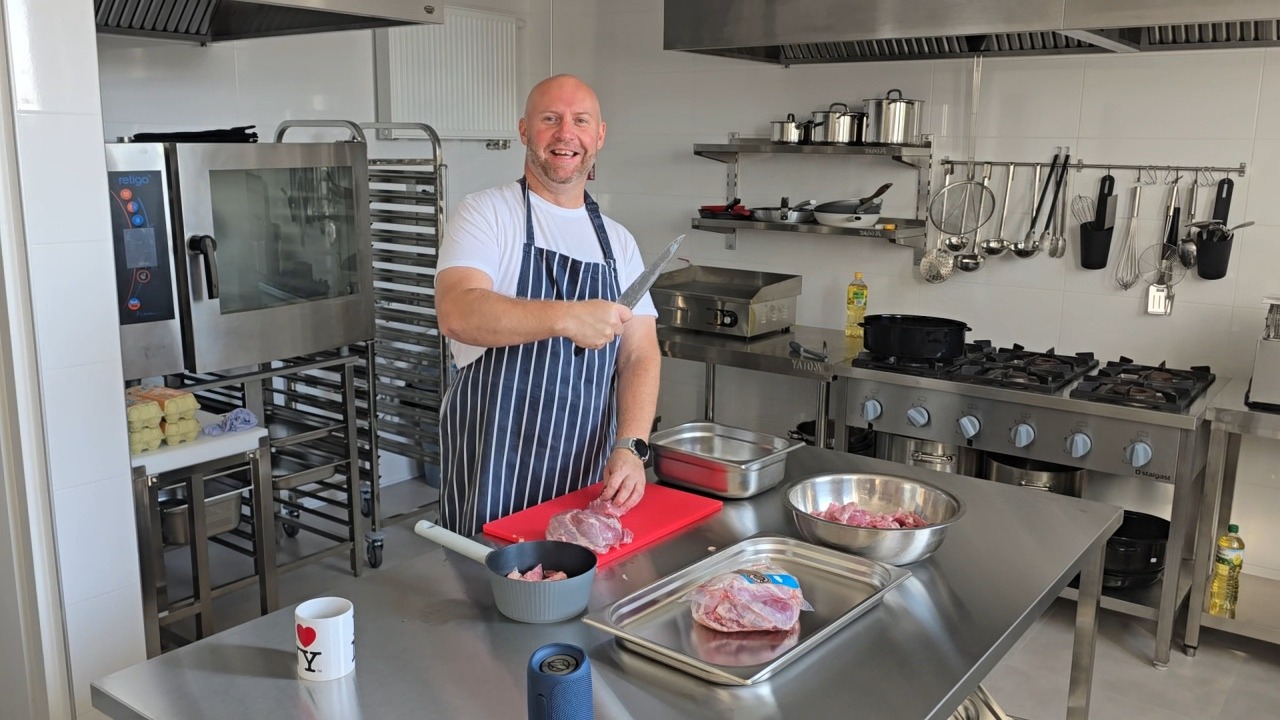 Kucharz w fartuchu kroi mięso na czerwonej desce w profesjonalnej kuchni. W tle piec konwekcyjny, stalowe półki z naczyniami i kuchenka gazowa. Na stole kubek z napisem.