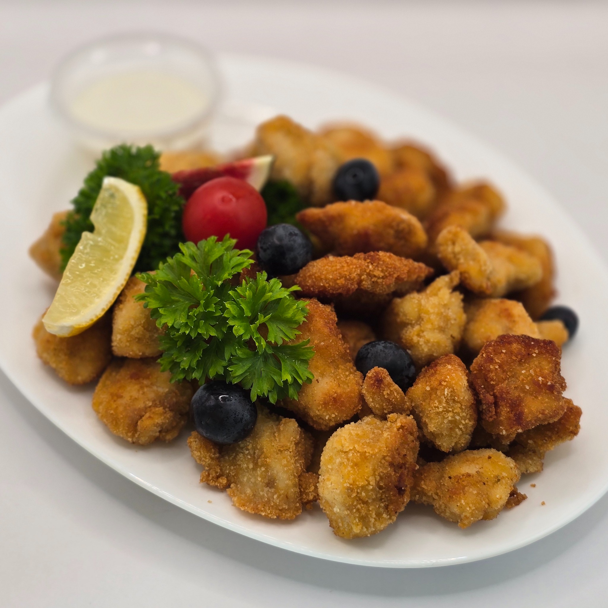 Apetyczne nuggetsy z kurczaka na białym talerzu, udekorowane świeżą natką pietruszki, borówkami, pomidorkiem cherry i cytryną, z sosem w tle. Idealne na catering.