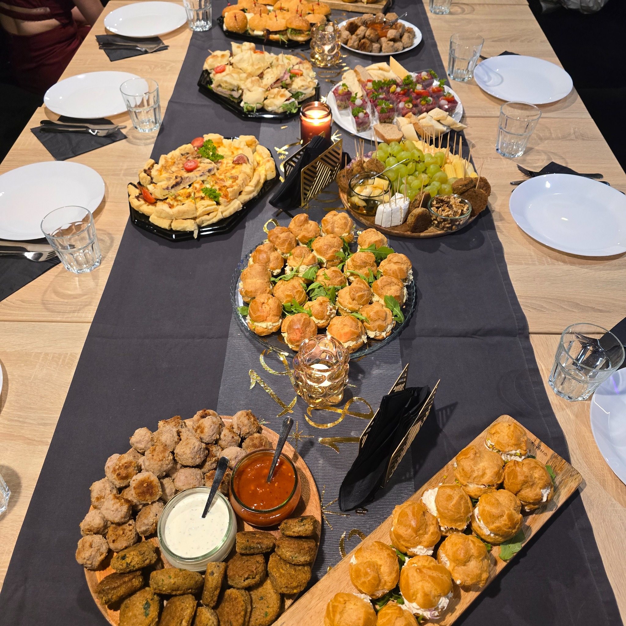 Stół cateringowy z różnorodnymi przekąskami: mini-burgery, tartaletki, koreczki, profiterolki, kulki mięsne i warzywne, sery, owoce. Dekoracja świecami i czarnymi serwetkami.