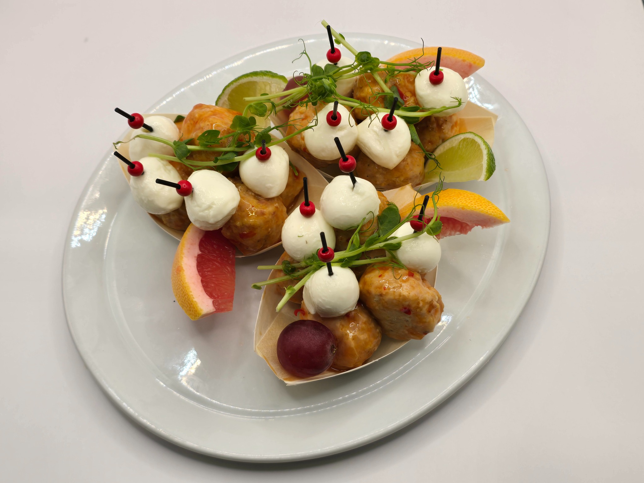 Eleganckie przekąski: mini-szaszłyki z kulek mozzarelli i mięsnych kulek, udekorowane cytrusami i mikro-ziołami, podane na białym talerzu. Idealne na catering.