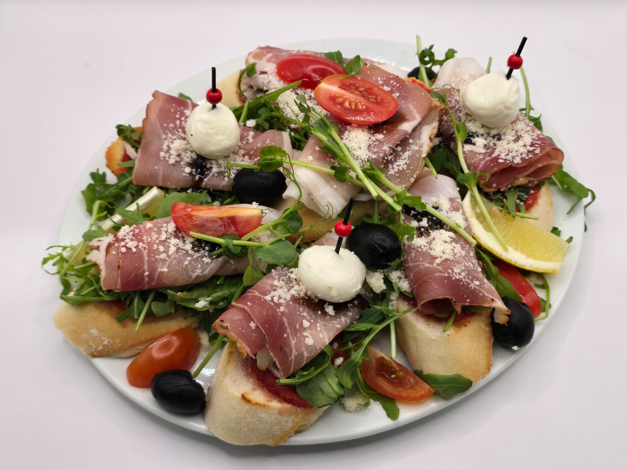 Apetyczna kompozycja: bruschetta z szynką parmeńską, pomidorkami, rukolą, mozzarellą na wykałaczkach i czarnymi oliwkami, posypana parmezanem, z cytryną na białym talerzu.