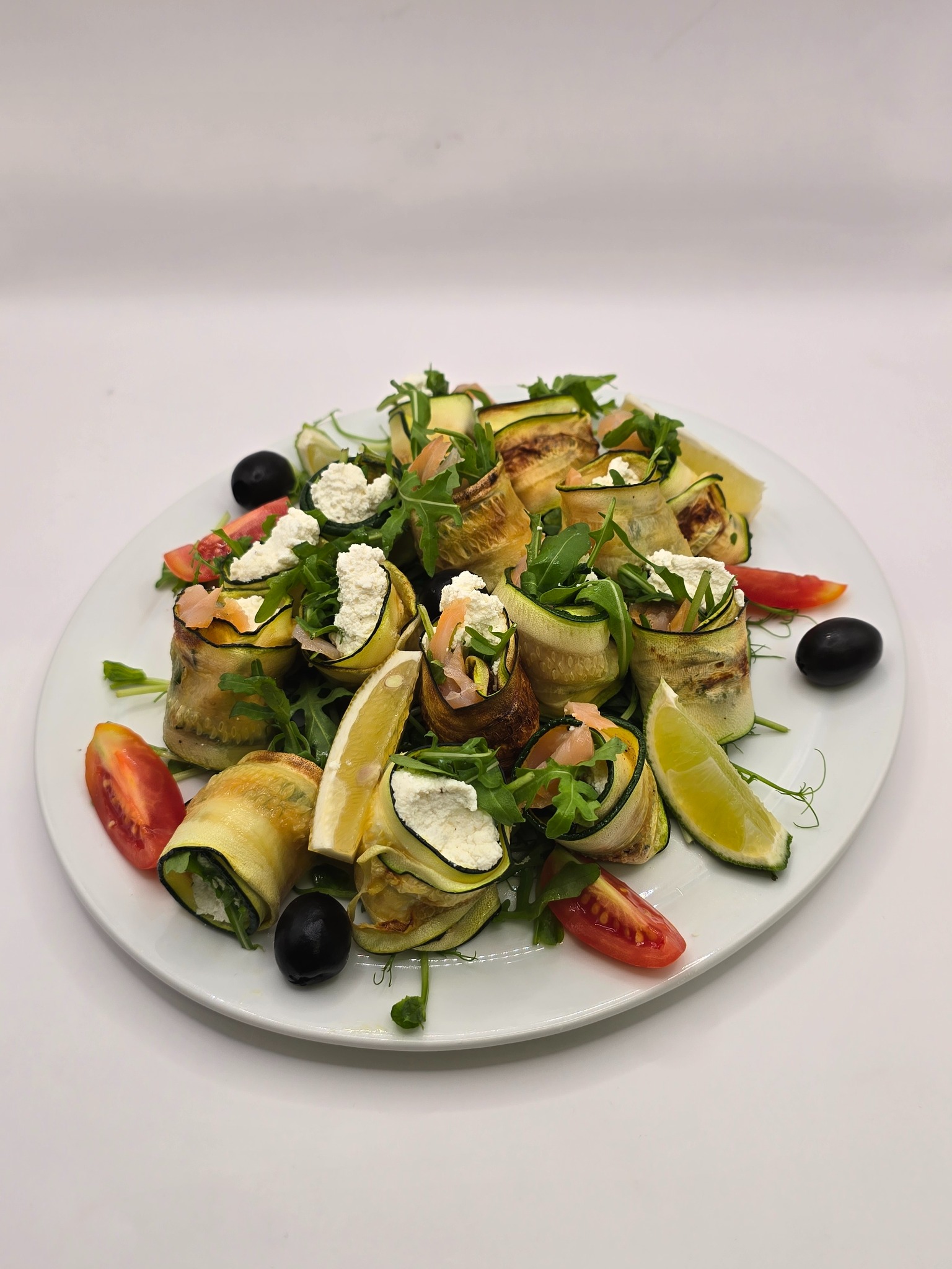 Elegancko podane roladki z cukinii z serem, rukolą, pomidorami i oliwkami na białym talerzu, udekorowane cytryną i limonką. Idealne na catering.