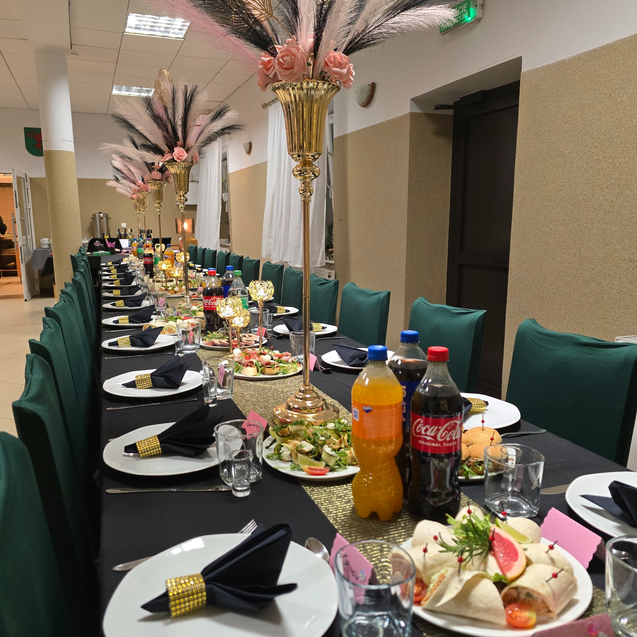 Uroczysty stół z czarnym obrusem, złotymi dekoracjami, kwiatami w wysokich wazonach i przekąskami. Przygotowany na imprezę firmową lub świąteczną.