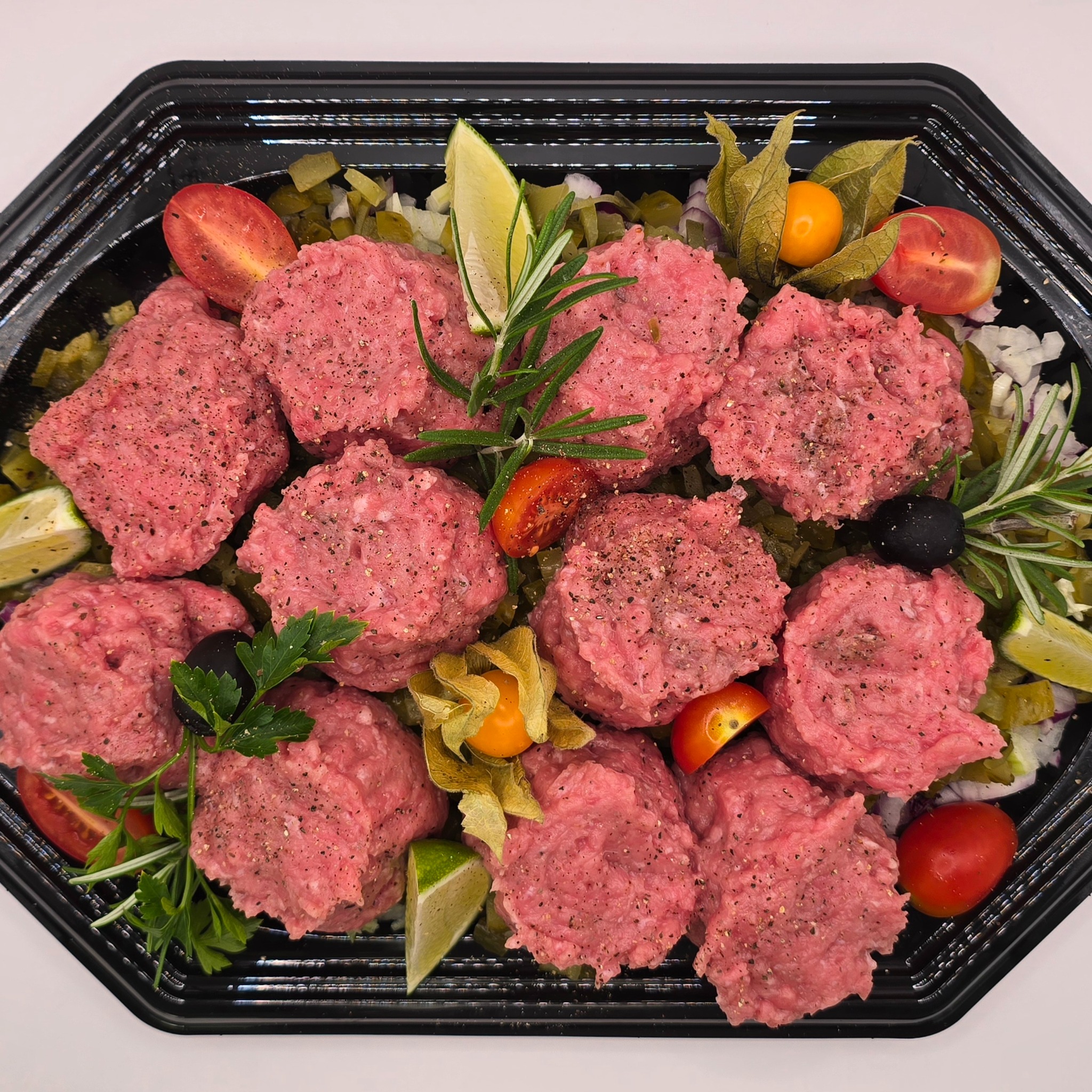 Czarny, ośmiokątny półmisek z tatarem wołowym, kaparami, ogórkiem, cebulą, pomidorkami koktajlowymi, oliwkami, physalis i ziołami, na białym tle.