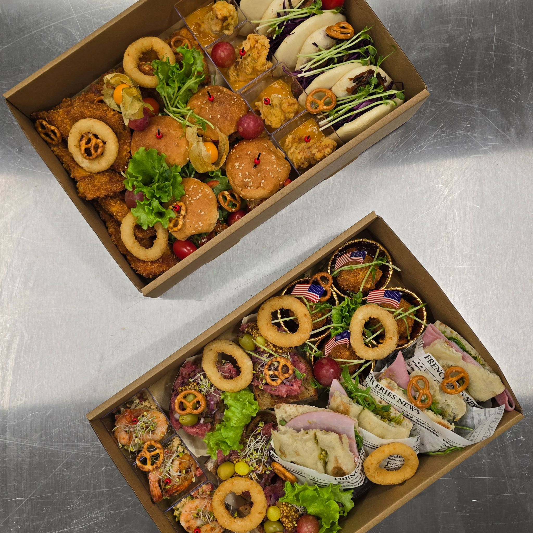 Dwie kartonowe tace z różnorodnymi przekąskami: mini burgery, krewetki, kanapki, krążki cebulowe i inne. Dekoracje z zieleniny i precelków. Idealne na imprezę firmową.