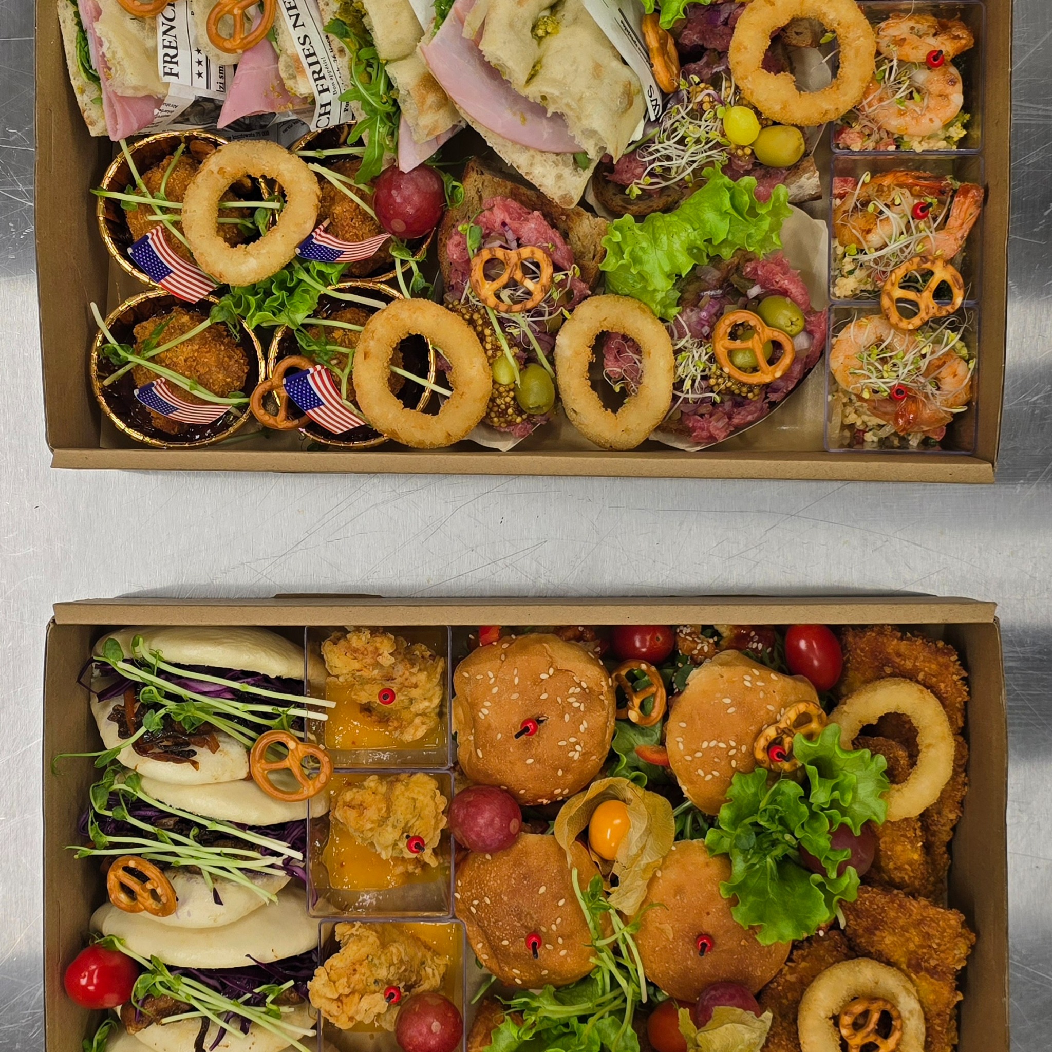Dwie kartonowe tace z różnorodnymi przekąskami: mini burgery, bao, krewetki, tartinki, krążki cebulowe i inne, udekorowane zieleniną i preclami.