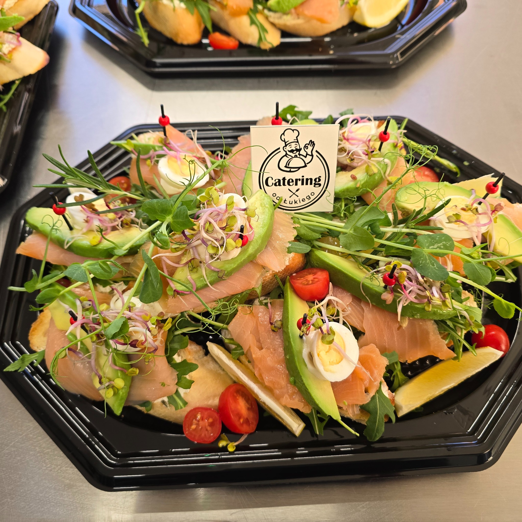 Elegancka taca cateringowa z kanapkami: łosoś, awokado, jajko przepiórcze, pomidorki koktajlowe i kiełki, ozdobiona logo firmy cateringowej.