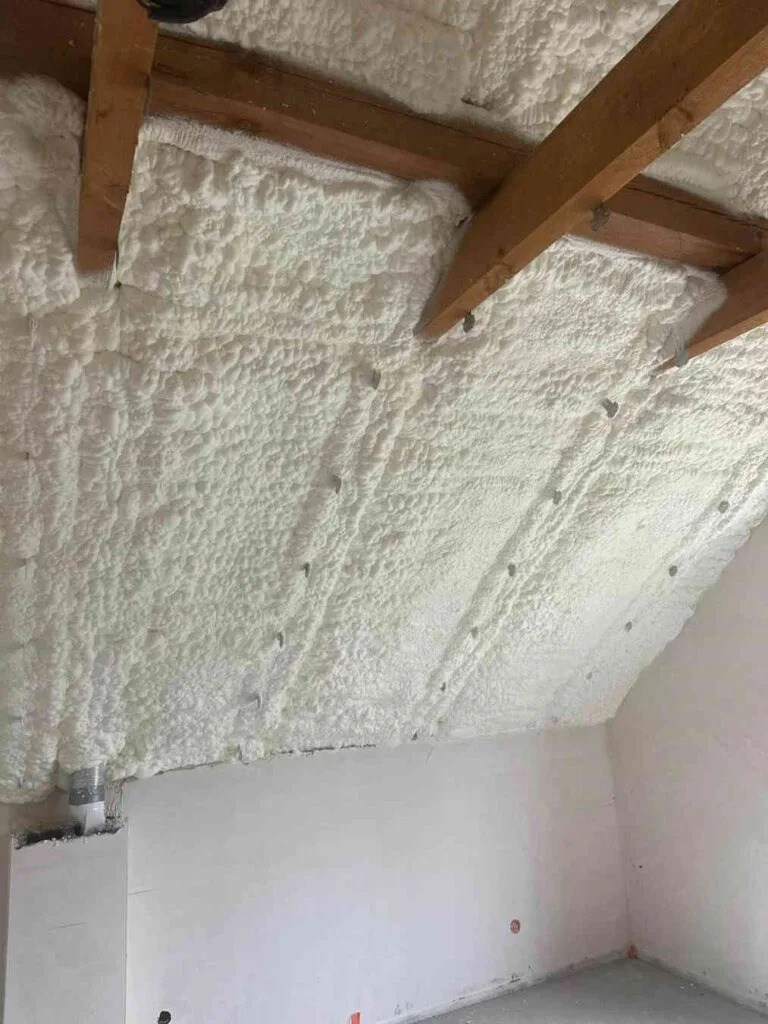 Poddasze po aplikacji pianki PUR, widoczne drewniane belki stropowe i biała, nierówna powierzchnia izolacji. Detal: szare elementy mocujące w izolacji.
