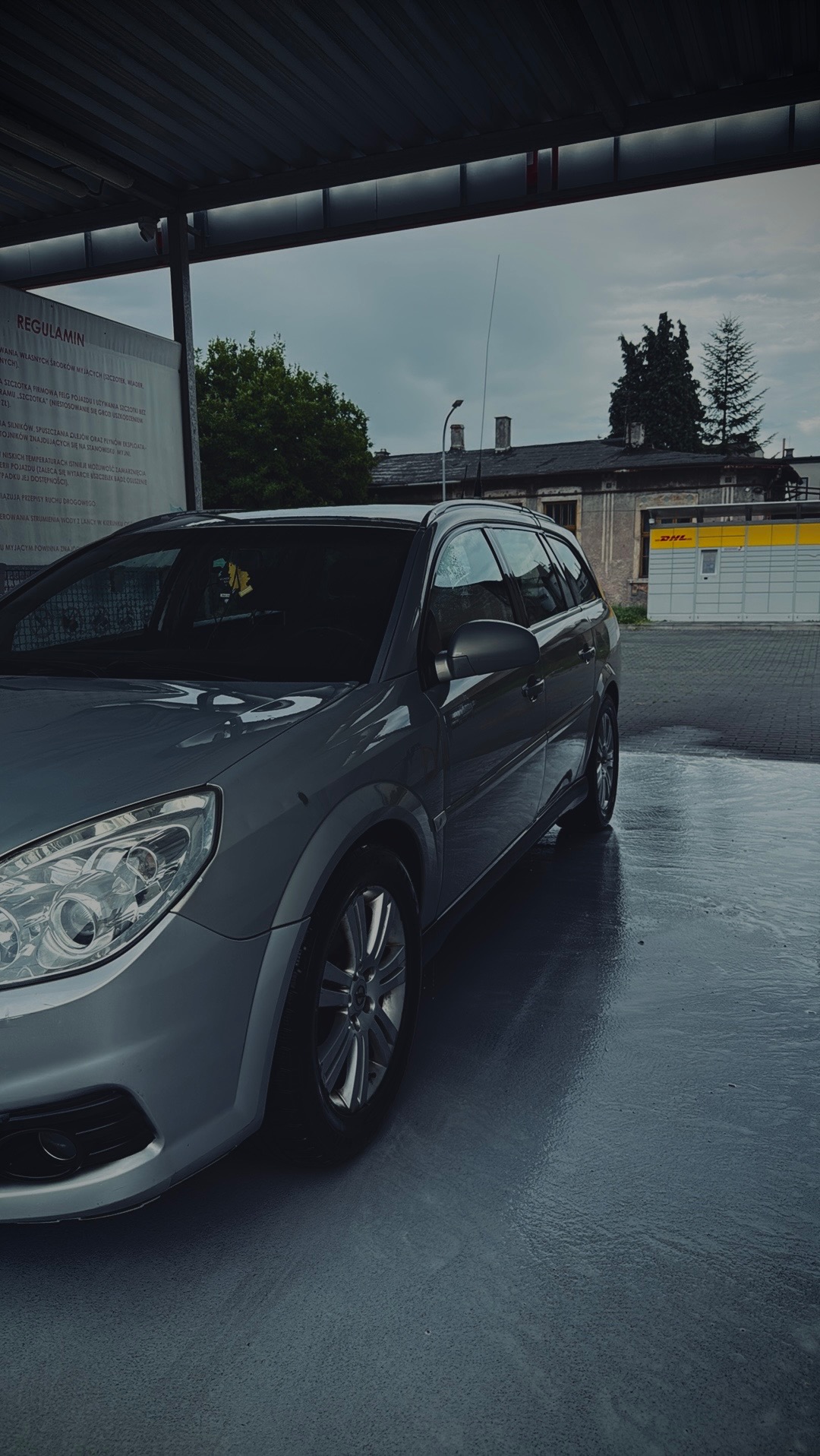 Szary samochód kombi po umyciu na myjni bezdotykowej, widoczny regulamin i budynek DHL w tle. Mokra nawierzchnia, pochmurne niebo.
