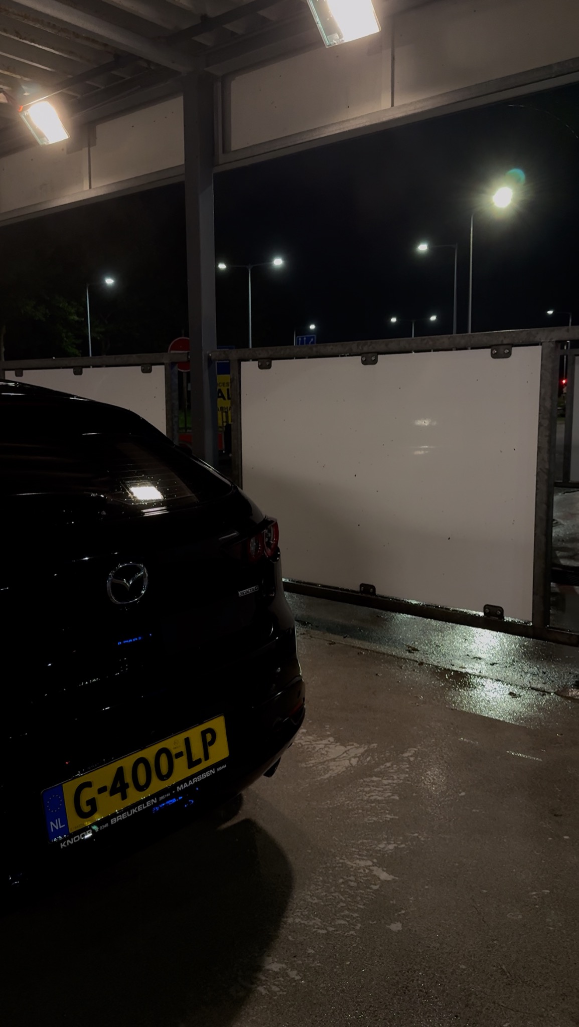 Częściowo widoczny, mokry, czarny samochód Mazda w myjni samochodowej nocą, z holenderską tablicą rejestracyjną G-400-LP.