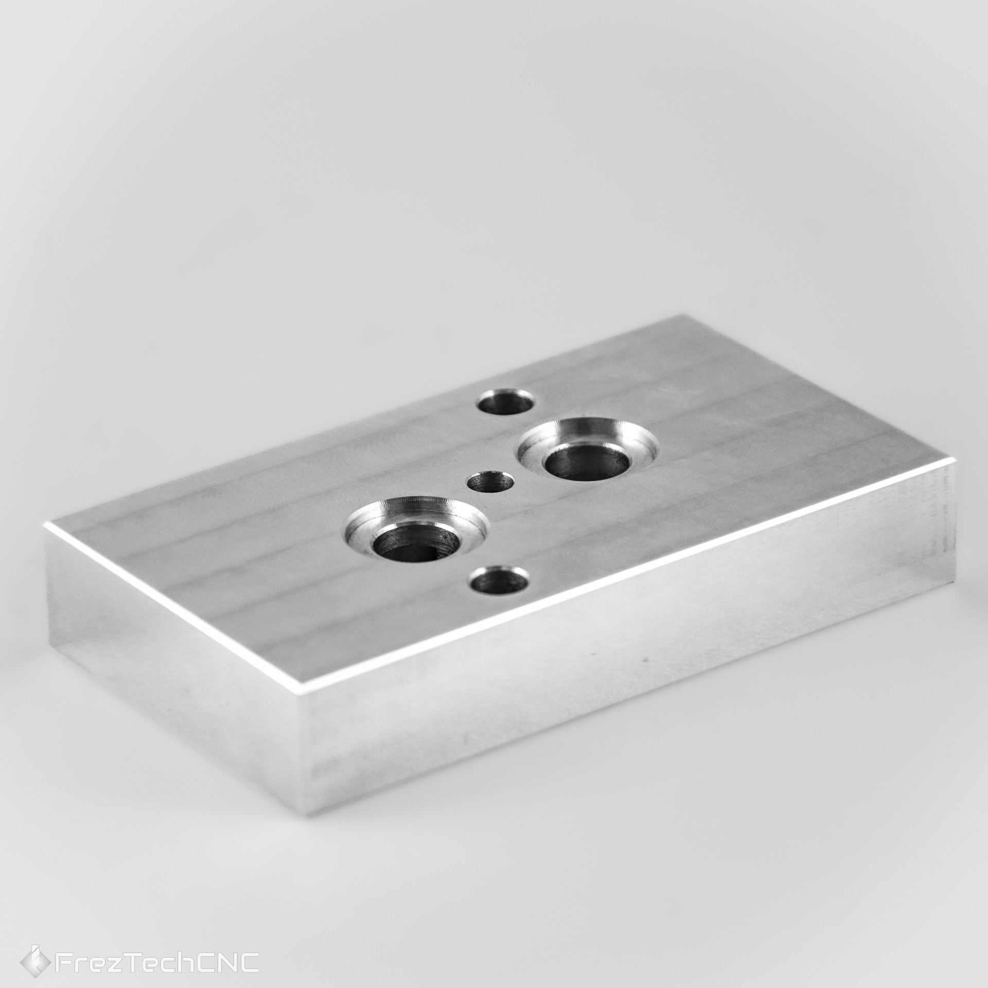 Precyzyjnie wykonany, srebrny element metalowy z otworami o różnych średnicach, obrobiony na maszynie. Widoczne ślady frezowania.