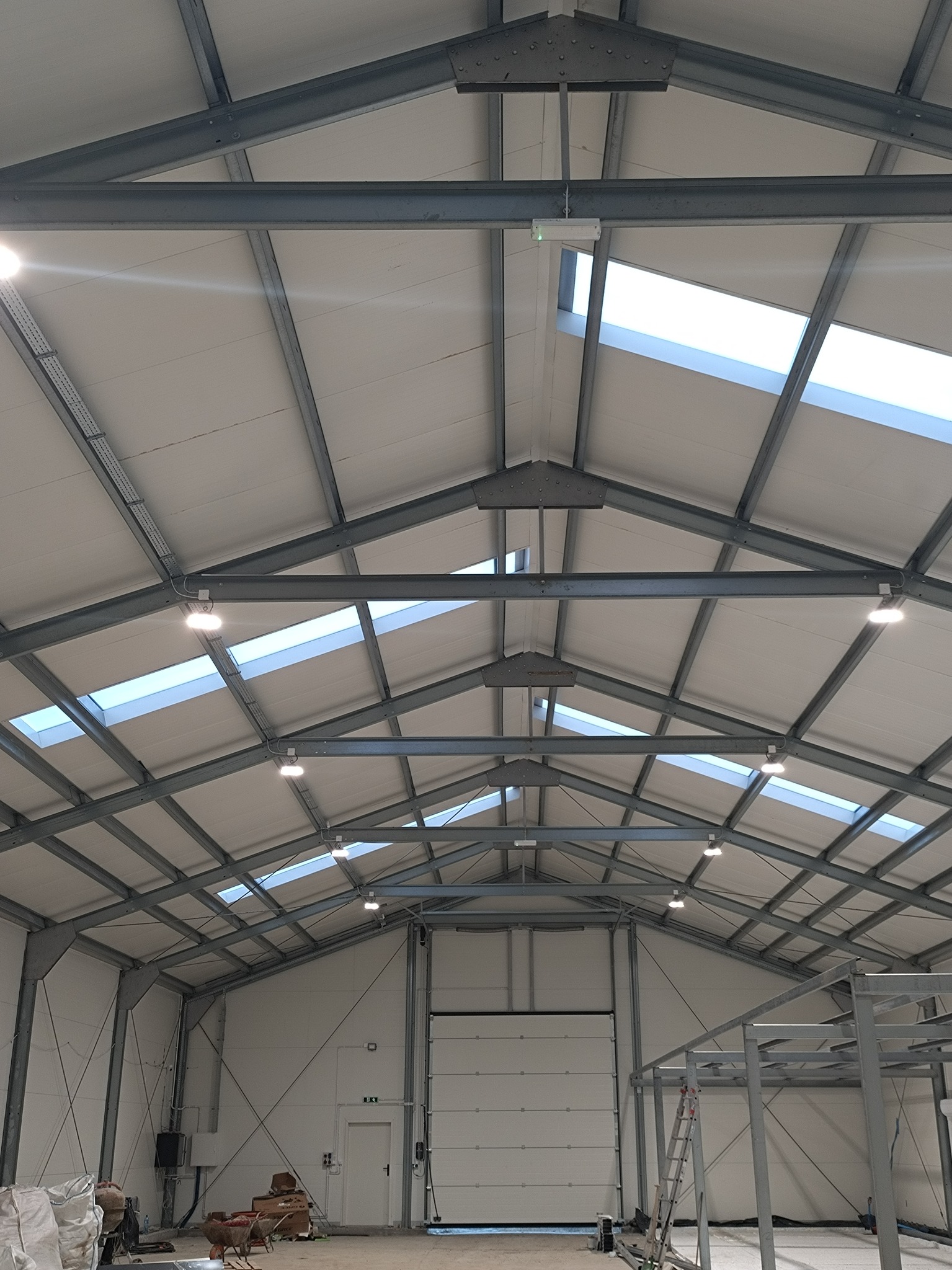 Wnętrze hali z metalową konstrukcją dachu, oświetleniem LED i świetlikami. Widoczne elementy instalacji elektrycznej i skrzynka elektryczna na ścianie.