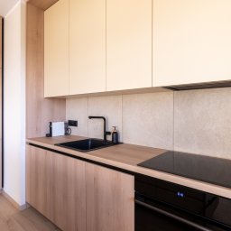 Bukabu Anna Pilch Projektowanie Wnętrz - Jasna kuchnia z drewnianymi szafkami i czarnym zlewem. Minimalistyczny design, beżowe płytki na ścianie, nowoczesna płyta indukcyjna. Oświetlenie górne.