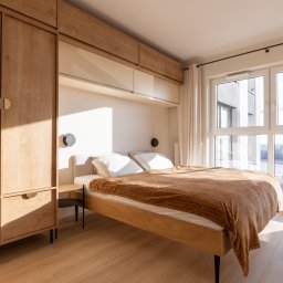 Bukabu Anna Pilch Projektowanie Wnętrz - Jasna sypialnia z drewnianą zabudową: szafa z półkami nad łóżkiem. Naturalne światło z okna oświetla parkiet i brązowy pled na łóżku. Minimalistyczny design.