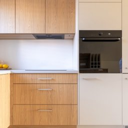 Bukabu Anna Pilch Projektowanie Wnętrz - Jasna kuchnia z drewnianymi frontami szafek i białymi elementami. Widoczny piekarnik w zabudowie. Minimalistyczny design, naturalne światło. Ujęcie na detale zabudowy kuchennej.