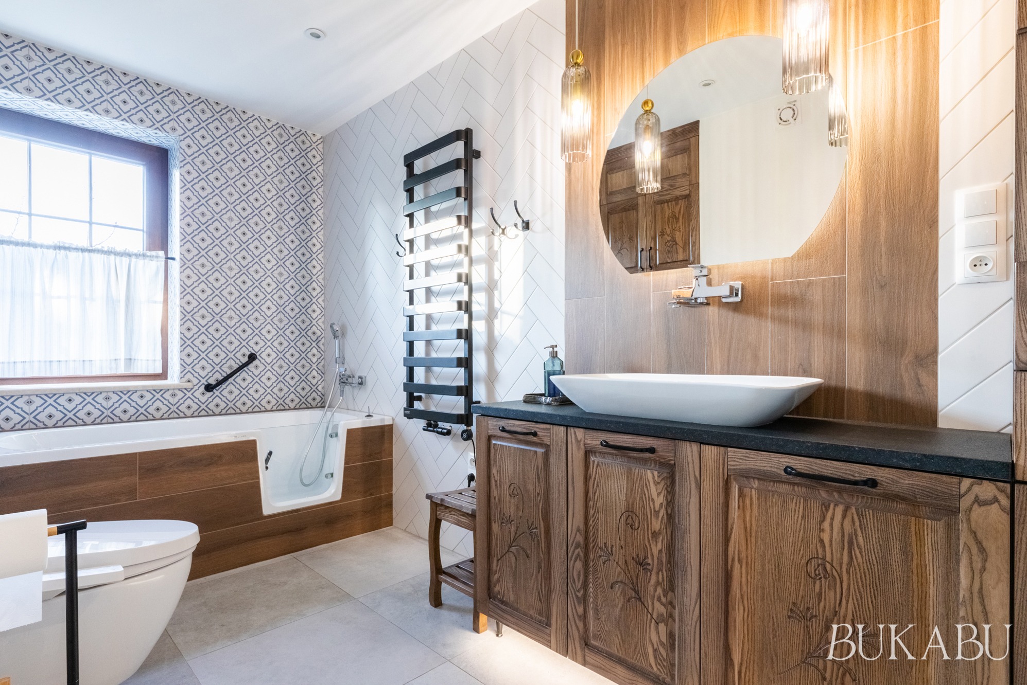 Funkcjonalna łazienka z wanną z drzwiczkami, umywalką nablatową i drewnianymi szafkami. Uwagę zwracają wzorzyste płytki i oświetlenie.