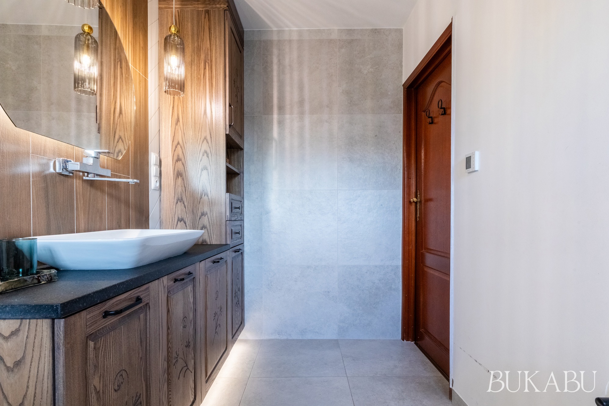 Nowoczesna łazienka z drewnianymi szafkami, owalną umywalką i szarą płytką. Wiszące lampy dodają elegancji. Widoczna część drzwi.