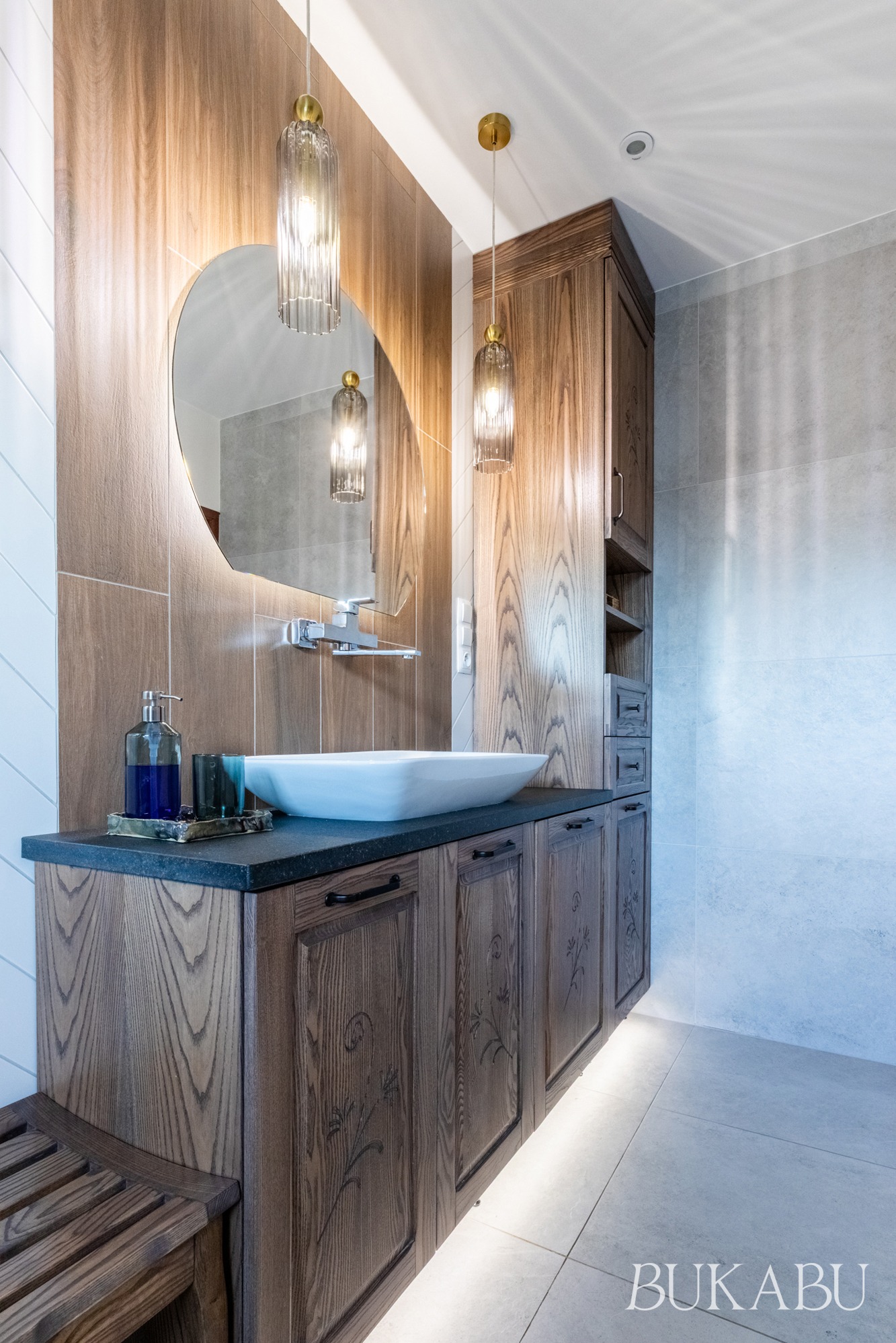 Nowoczesna łazienka z drewnianymi szafkami, owalnym lustrem z podświetleniem i wiszącymi lampami. Umywalka nablatowa, ciemny blat i subtelne oświetlenie pod szafkami.