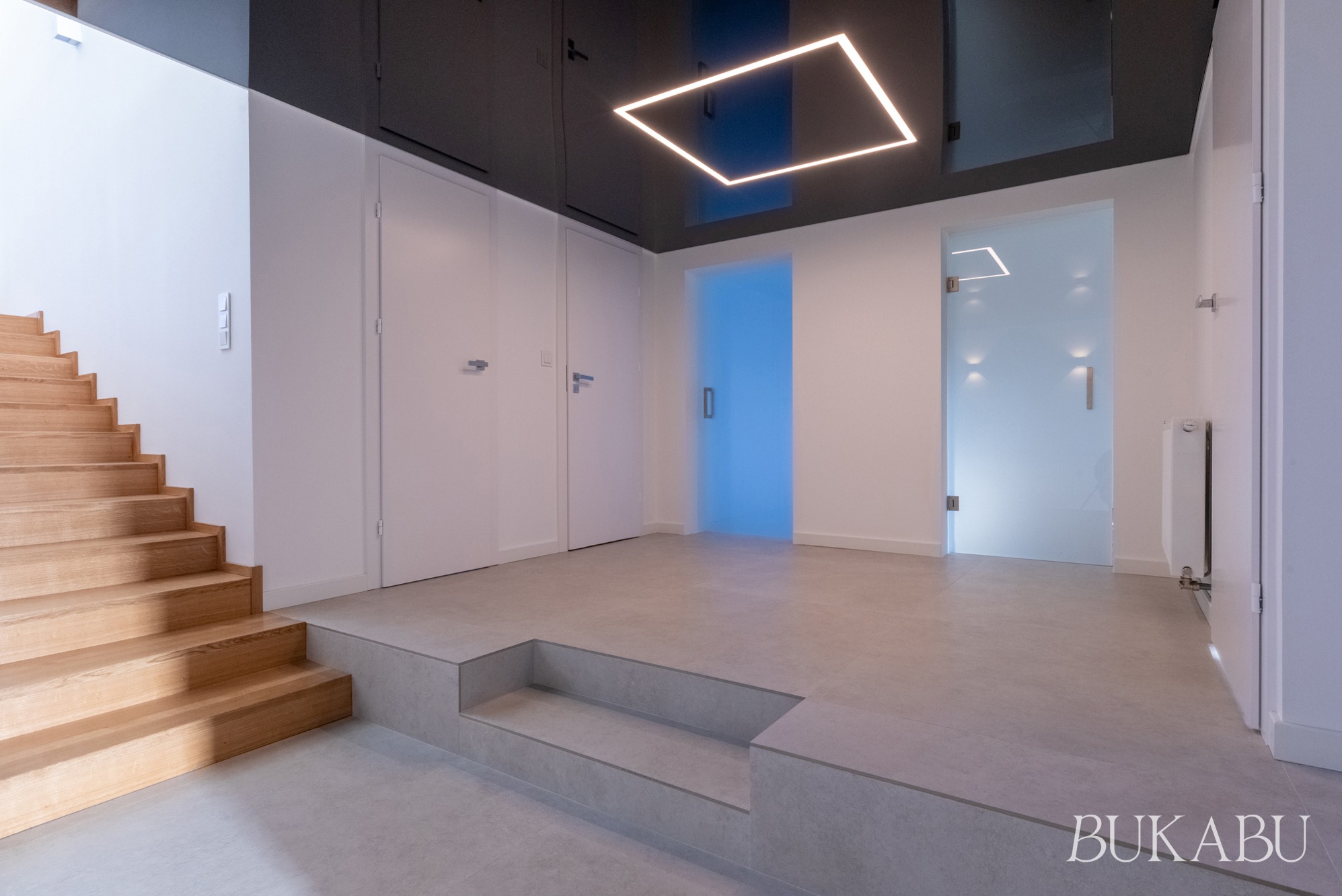 Nowoczesne wnętrze: drewniane schody, szara podłoga, białe ściany i drzwi, podwieszany sufit z kwadratową lampą LED. Minimalistyczny design z akcentem kolorystycznym.
