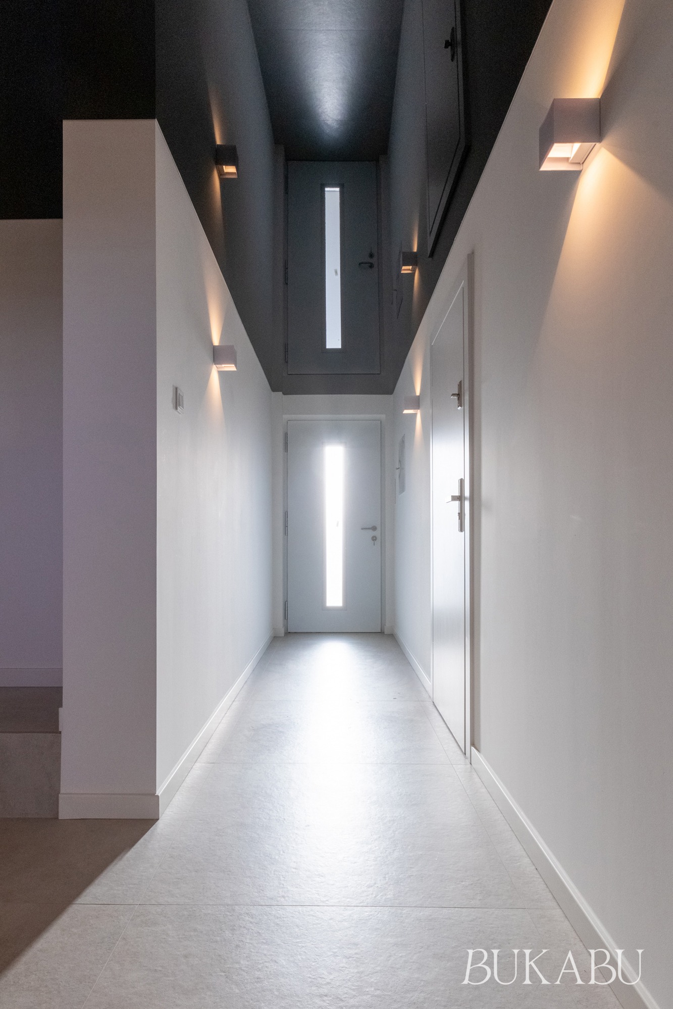 Nowoczesny korytarz w bieli i szarości z minimalistycznym oświetleniem ściennym. Widok na drzwi wejściowe z przeszkleniem. Ciemny sufit z wbudowanymi lampami.