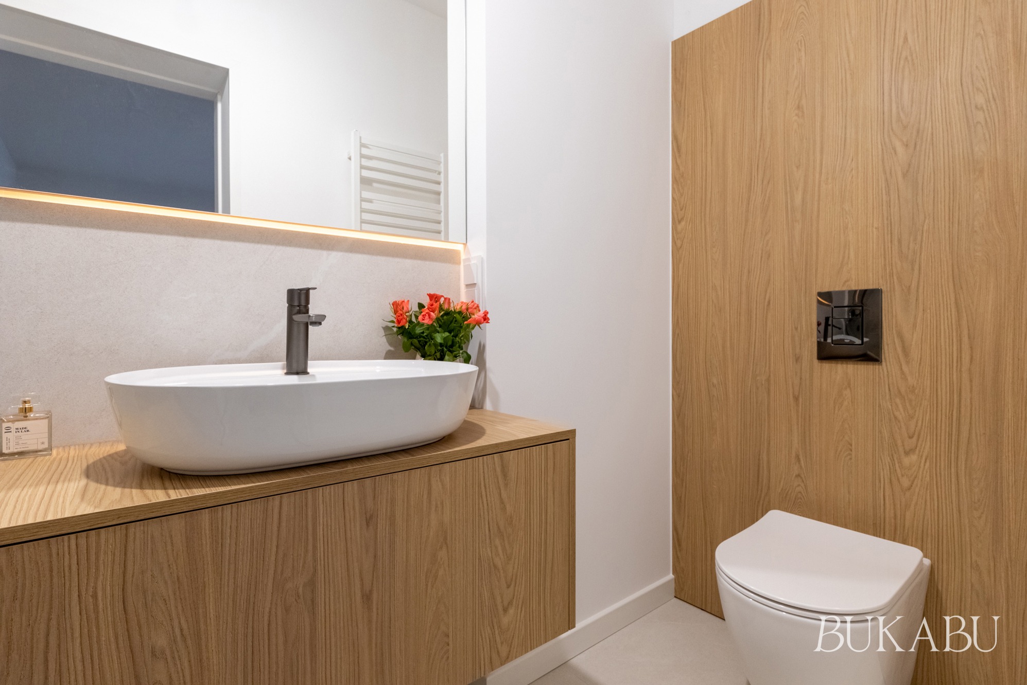 Nowoczesna łazienka z drewnianymi akcentami: umywalka nablatowa, lustro z podświetleniem LED, toaleta w zabudowie. Minimalistyczny design, ciepłe oświetlenie.