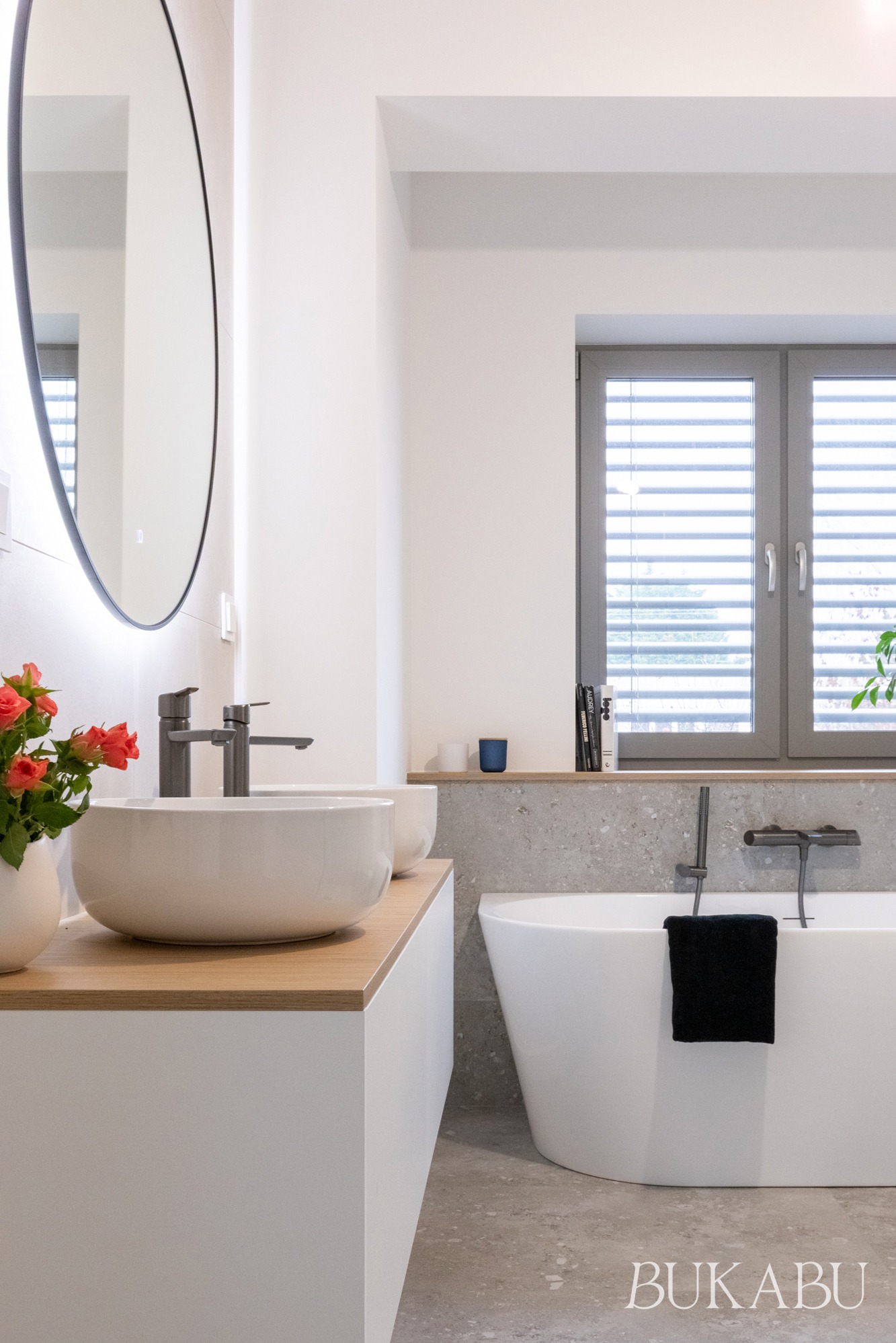 Jasna łazienka z owalną umywalką na drewnianym blacie, okrągłym lustrem z podświetleniem i wolnostojącą wanną przy oknie z żaluzjami. Minimalistyczny design.