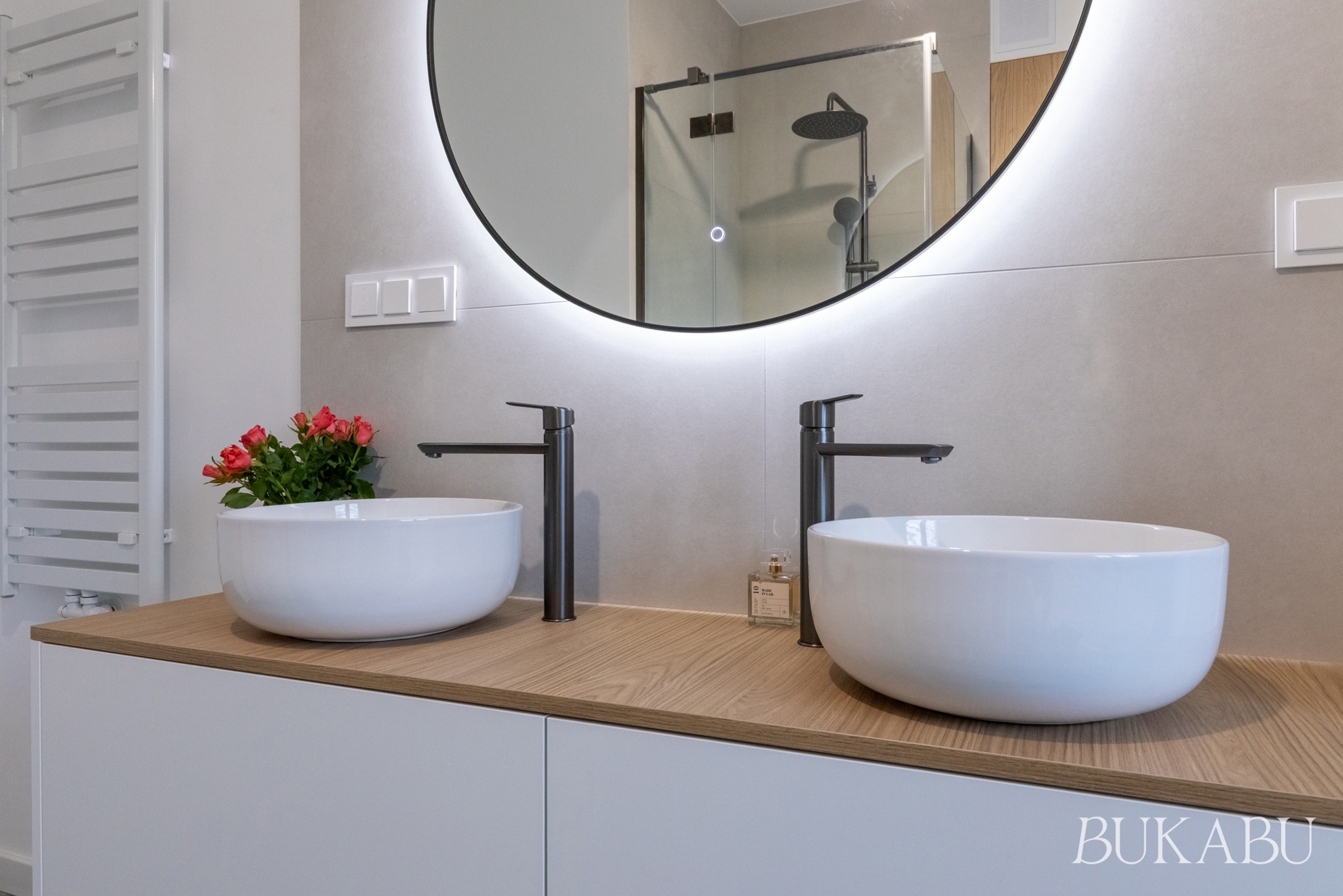 Nowoczesna łazienka z dwoma białymi umywalkami na drewnianym blacie, czarne baterie, okrągłe lustro z podświetleniem LED i szklana kabina prysznicowa w tle.