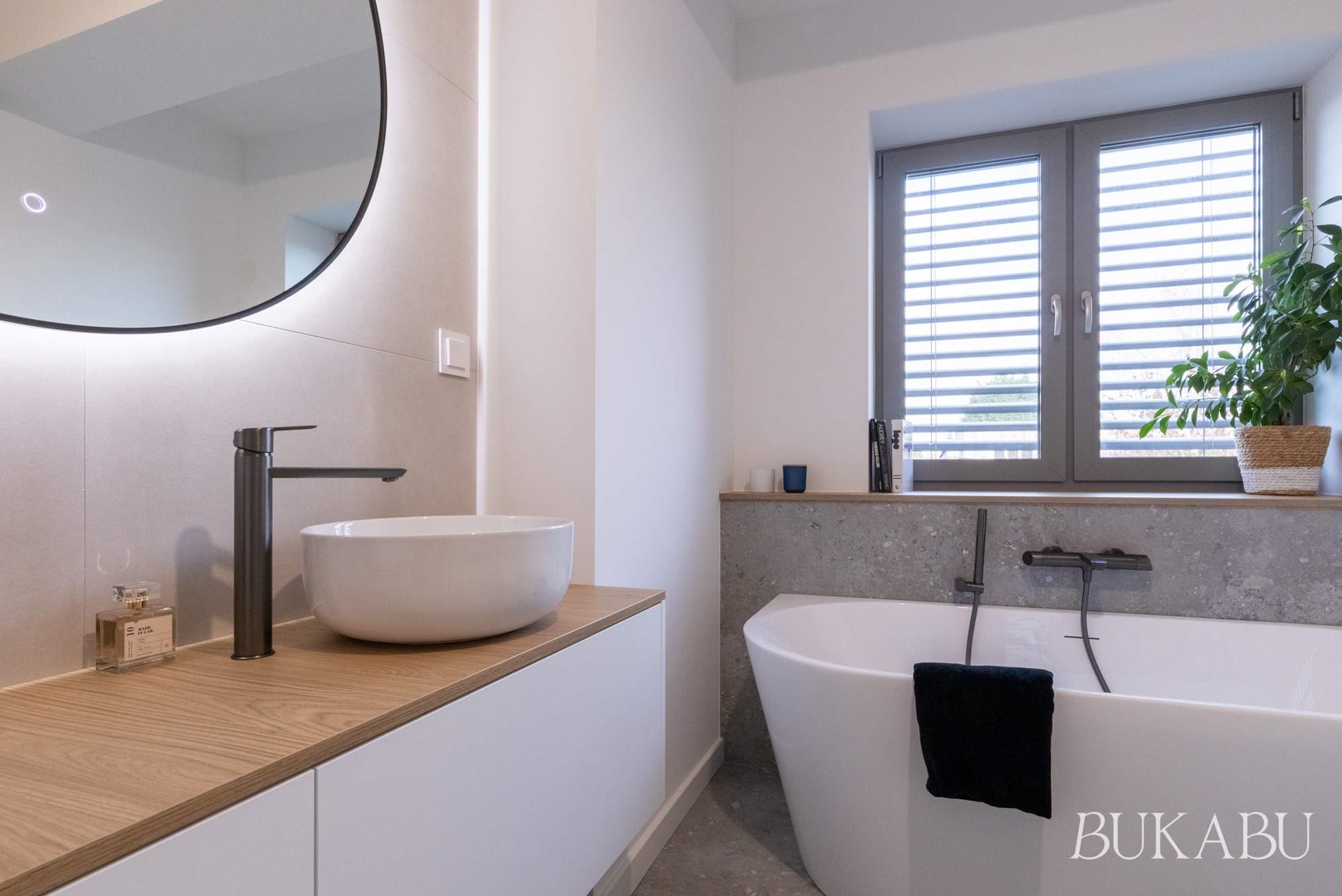 Nowoczesna łazienka z okrągłym lustrem z podświetleniem LED, białą umywalką nablatową i wolnostojącą wanną przy oknie z żaluzjami. Minimalistyczny design.