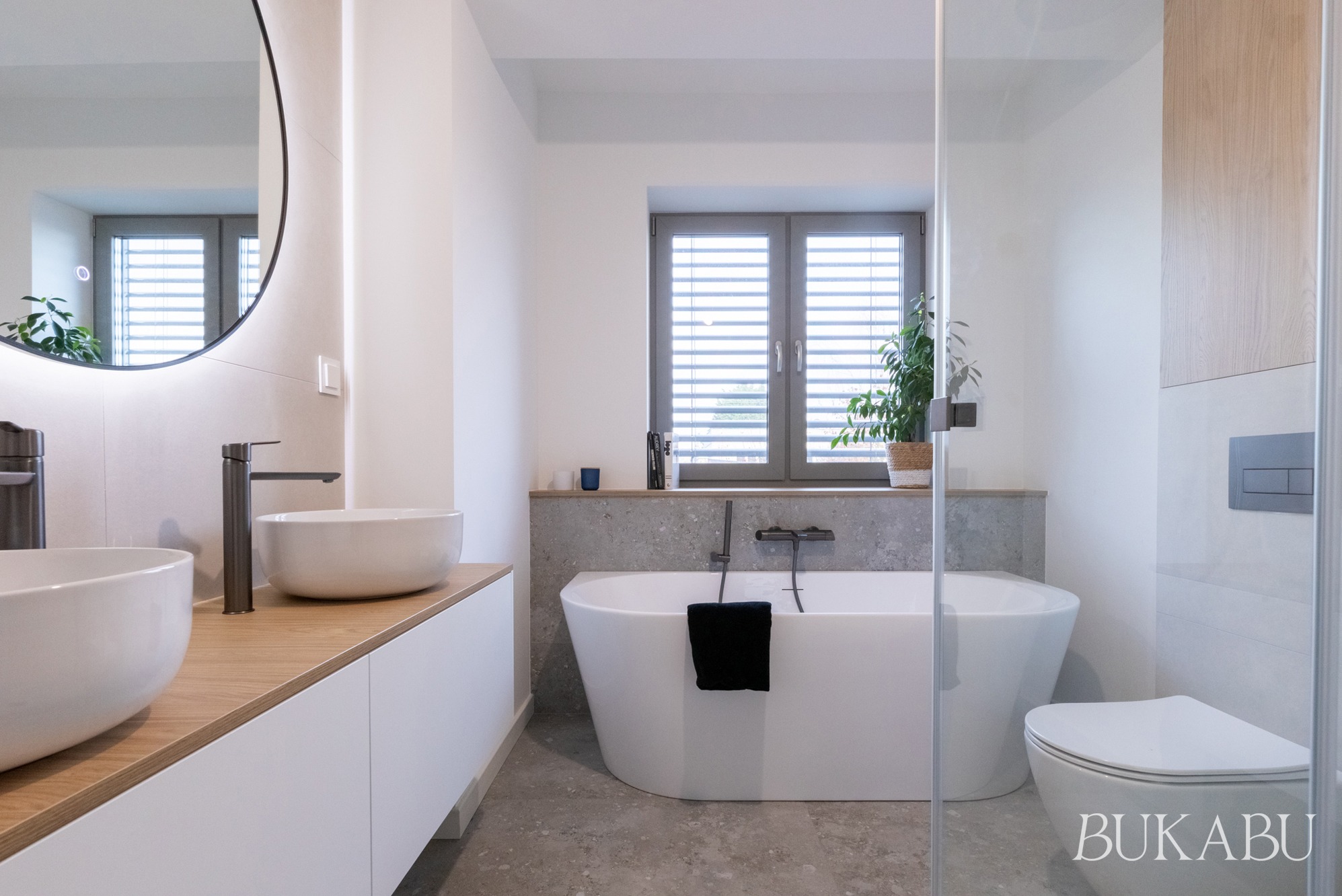Jasna łazienka z wanną wolnostojącą, umywalkami nablatowymi i dużym lustrem z podświetleniem. Minimalistyczny design z akcentami drewna i szarości.