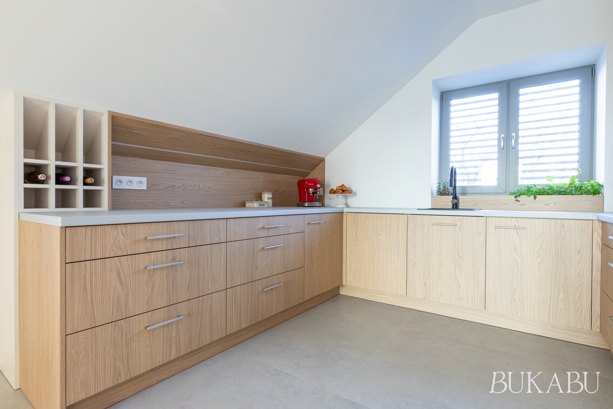 Jasna kuchnia na poddaszu z drewnianymi szafkami, szarym blatem i oknem z ziołami. Uwagę przyciąga czerwony ekspres do kawy i winiarka z butelkami wina.